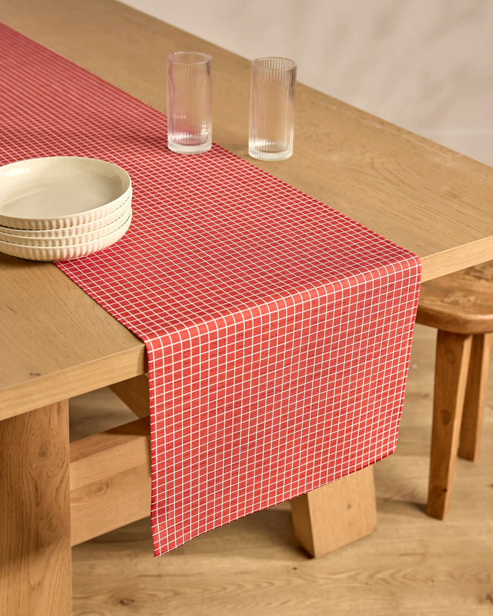 A light, natural-finish wooden dining table with the Candy Cane Grid table runner by Kailee Wright in red and white checks, two clear ribbed glasses, two stacked white bowls, and paired with wooden stools.