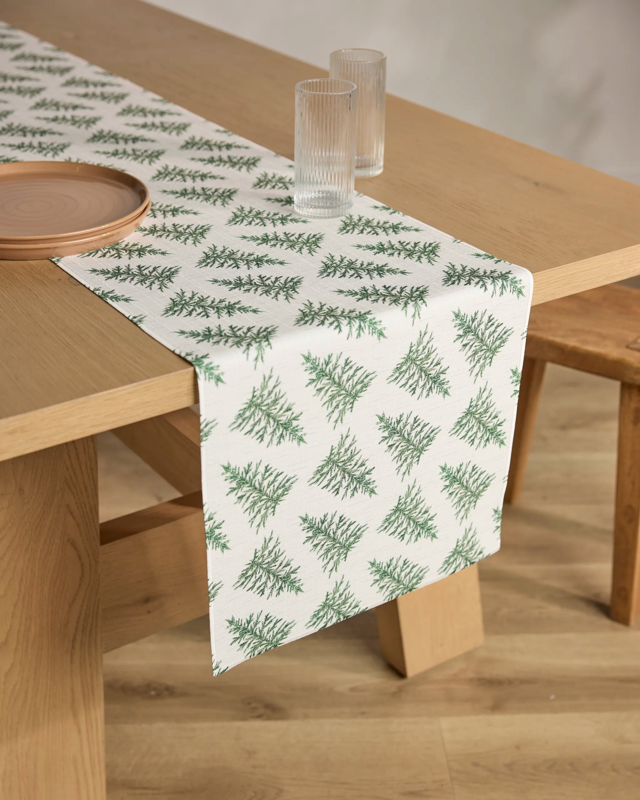 A Cedar wooden dining table by Geometry with a stain-resistant pine tree runner, two ribbed glass tumblers, a round ceramic plate, and a wooden bench on a light wood floor.
