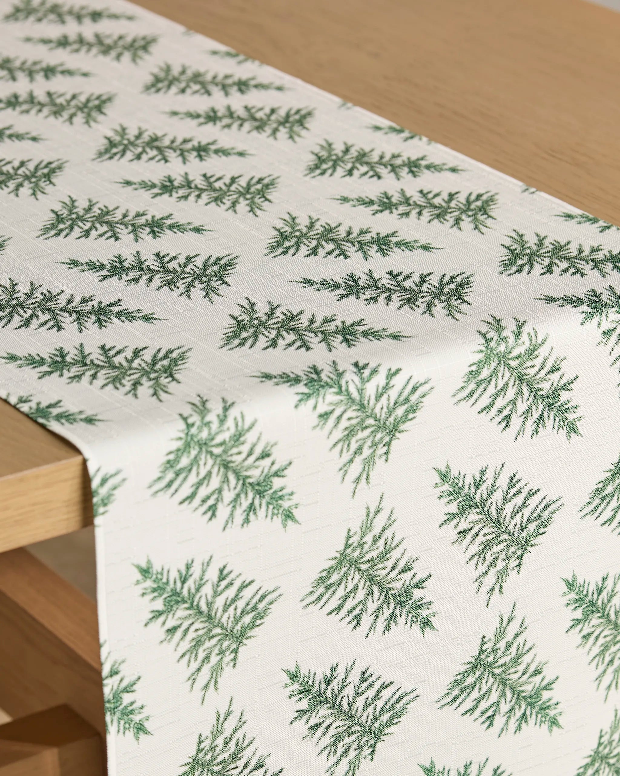 The Cedar table runner by Geometry, featuring a repeating pattern of green pine branches and stain-resistant fabric, is spread over a light wooden table.
