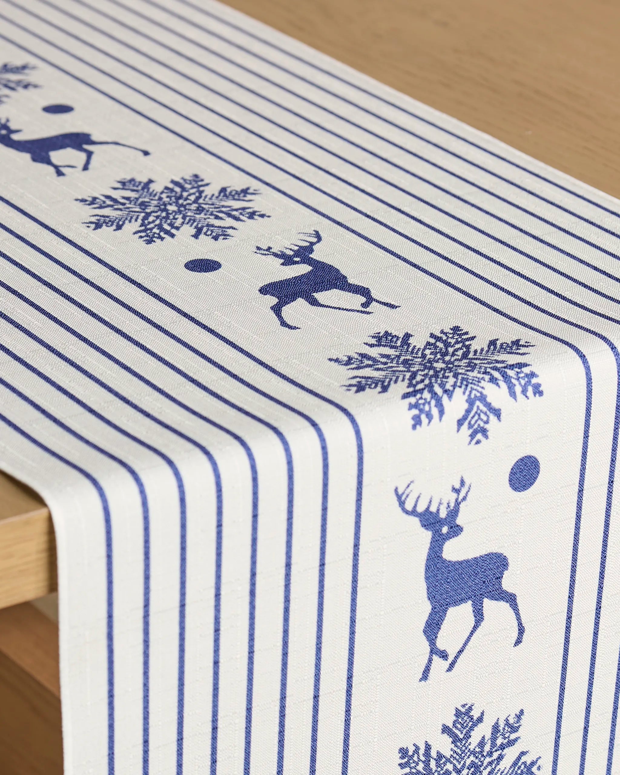 The Geometry Nordic Stag table runner features blue vertical stripes and blue silhouettes of reindeer, snowflakes, and dots, all stain-resistant and draped over a light wooden table.