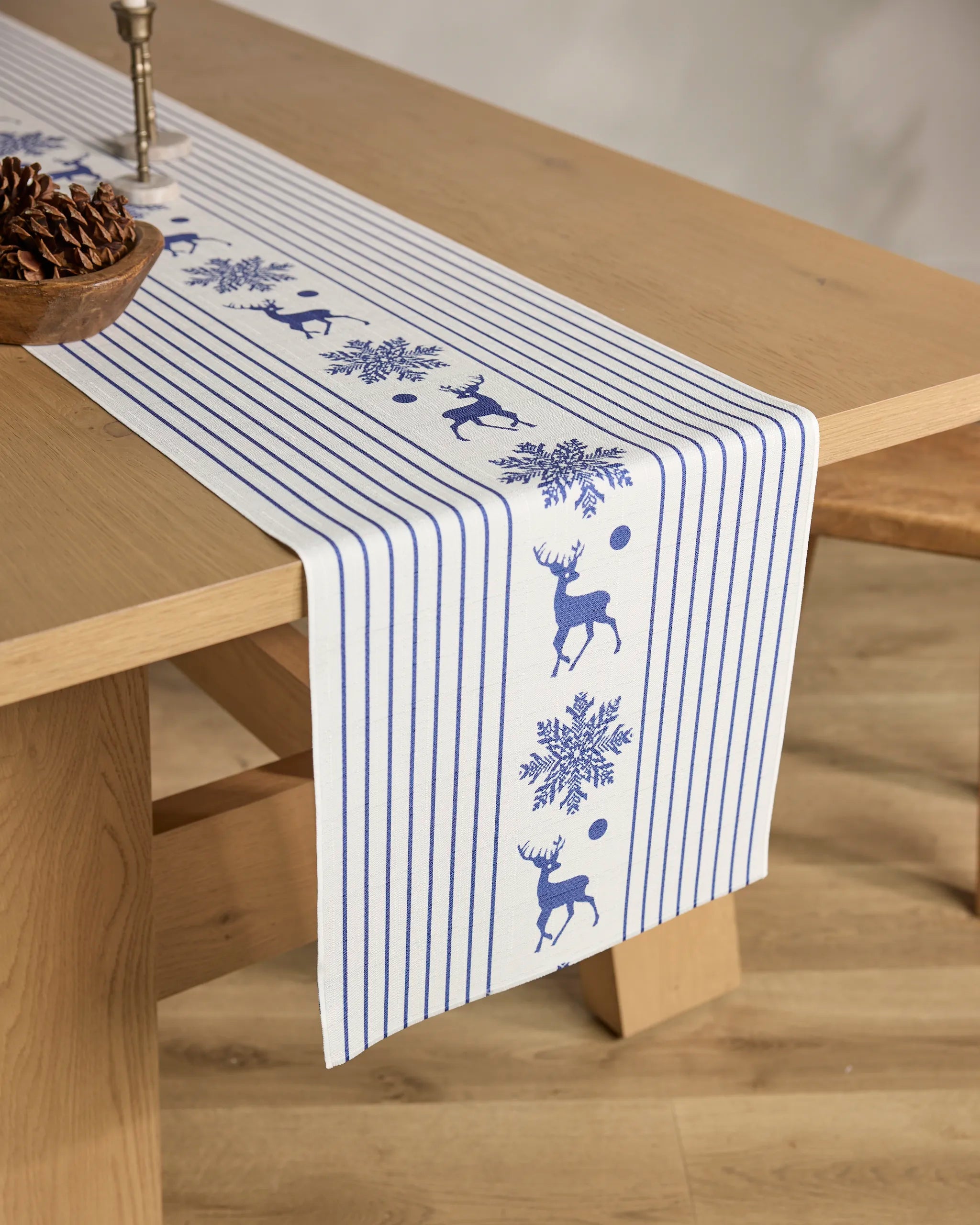 The Geometry Nordic Stag table runner features a blue and white striped, stain-resistant design with blue reindeer and snowflake motifs on a wooden table, partially topped with a pinecone bowl and candle.