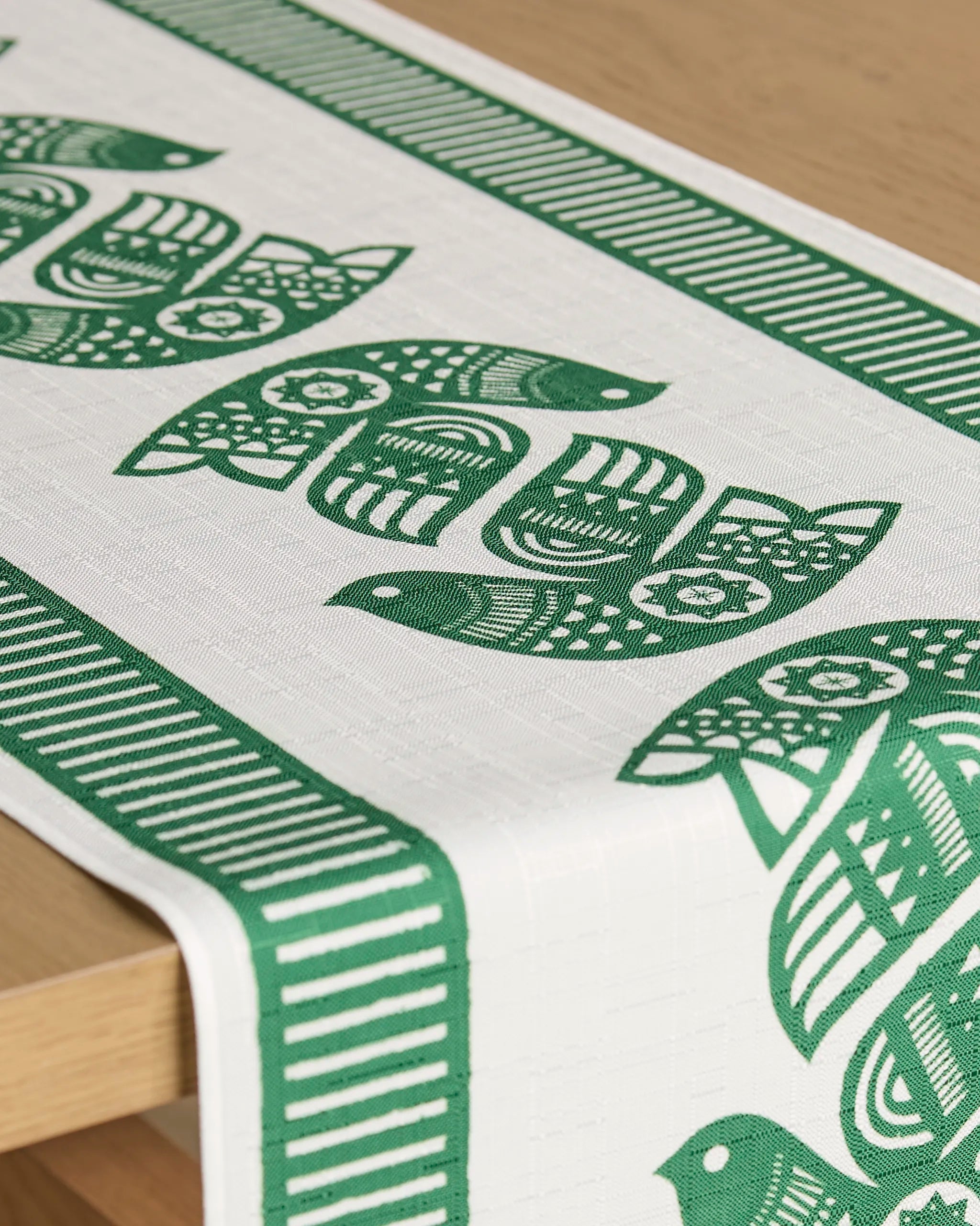 Geometry's "Peacebound" is a white, stain-resistant table runner with green patterned bird designs and green-striped borders, displayed draped over the corner of a light wooden table.