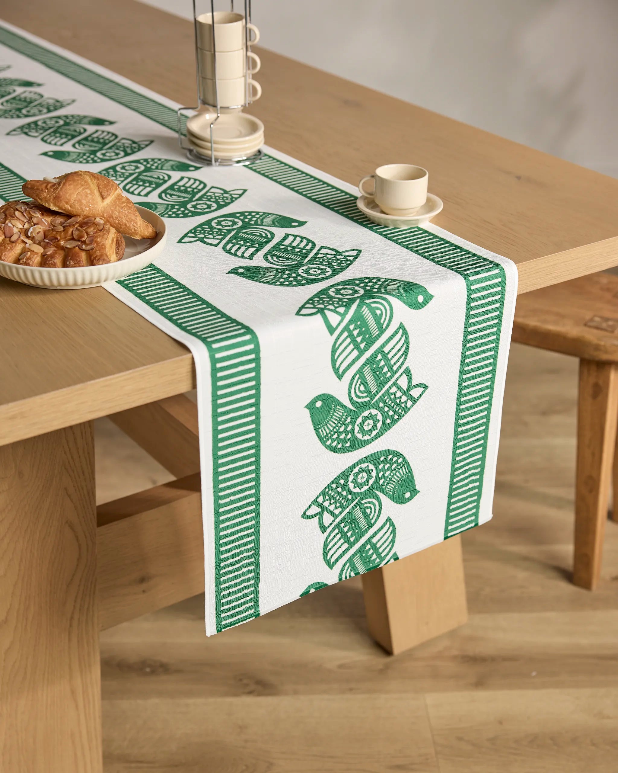 A wooden table topped with the Geometry Peacebound green and white stain-resistant table runner, featuring stylized birds, is set with a plate of pastries, cup and saucer, and pepper mill in a bright, cozy room.