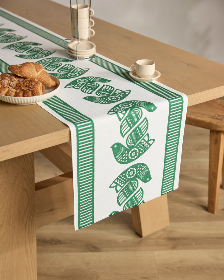 A wooden table topped with the Geometry Peacebound green and white stain-resistant table runner, featuring stylized birds, is set with a plate of pastries, cup and saucer, and pepper mill in a bright, cozy room.