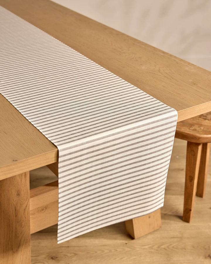 A light wooden table features the Kailee Wright Stone Stripe stain-resistant white and gray striped table runner, paired with matching wooden chairs on a light wood floor.