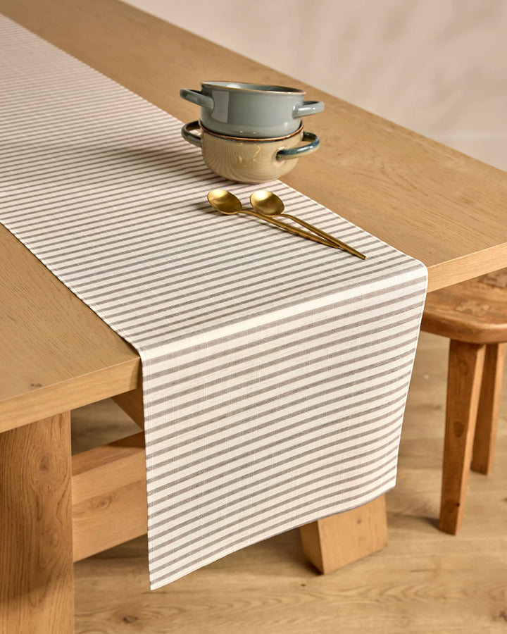 A light wooden table features the Kailee Wright Stone Stripe gray and white runner, two stacked blue-gray bowls on a matching saucer, and two gold spoons. Wooden stools and a wooden floor complete the setting.