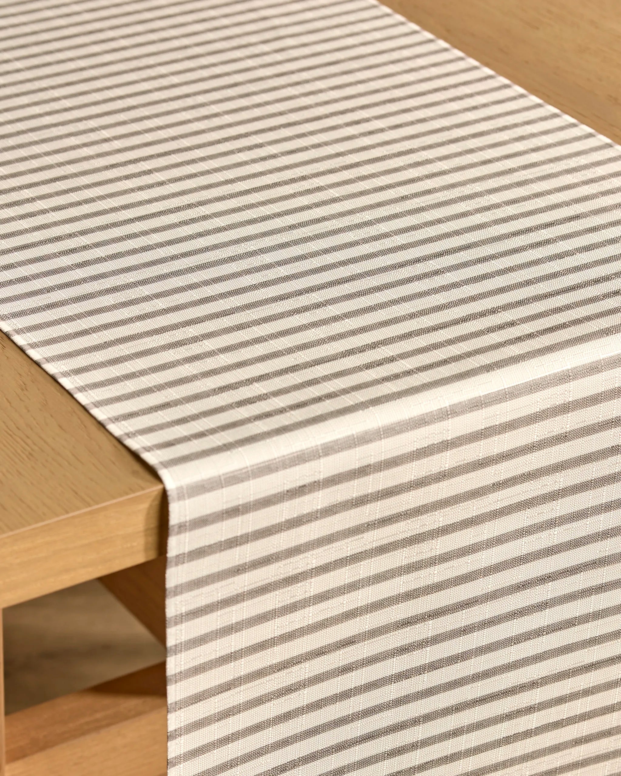 A close-up of a light wooden table with the Kailee Wright Stone Stripe table runner. Its beige and gray stripes form a subtle plaid pattern, combining style and practicality with stain-resistant fabric.