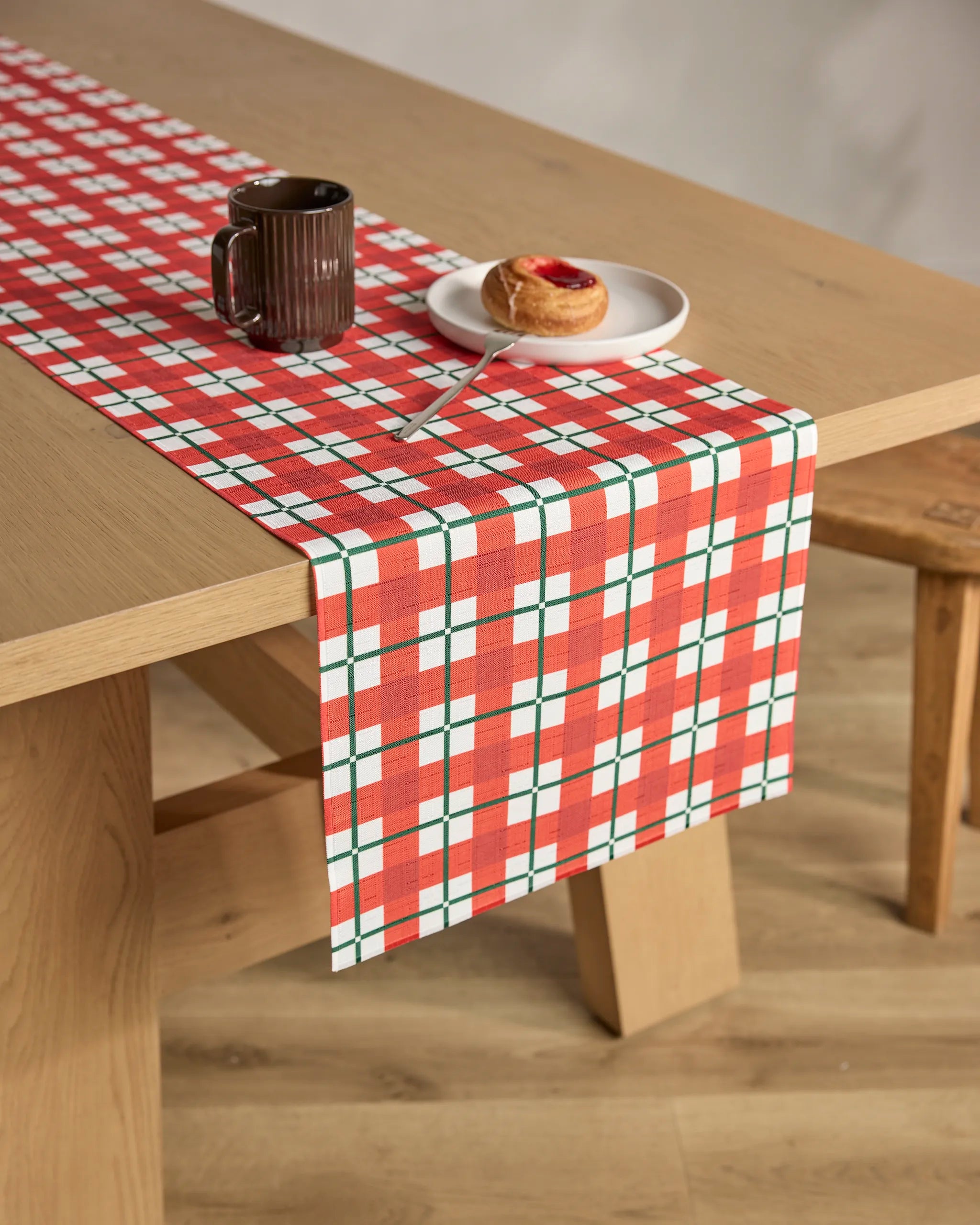 A wooden table topped with the Geometry Yuletide Plaid stain-resistant table runner holds a dark mug, white plate, jam pastry, and spoon. Next to it is a wooden bench on a light wood floor.