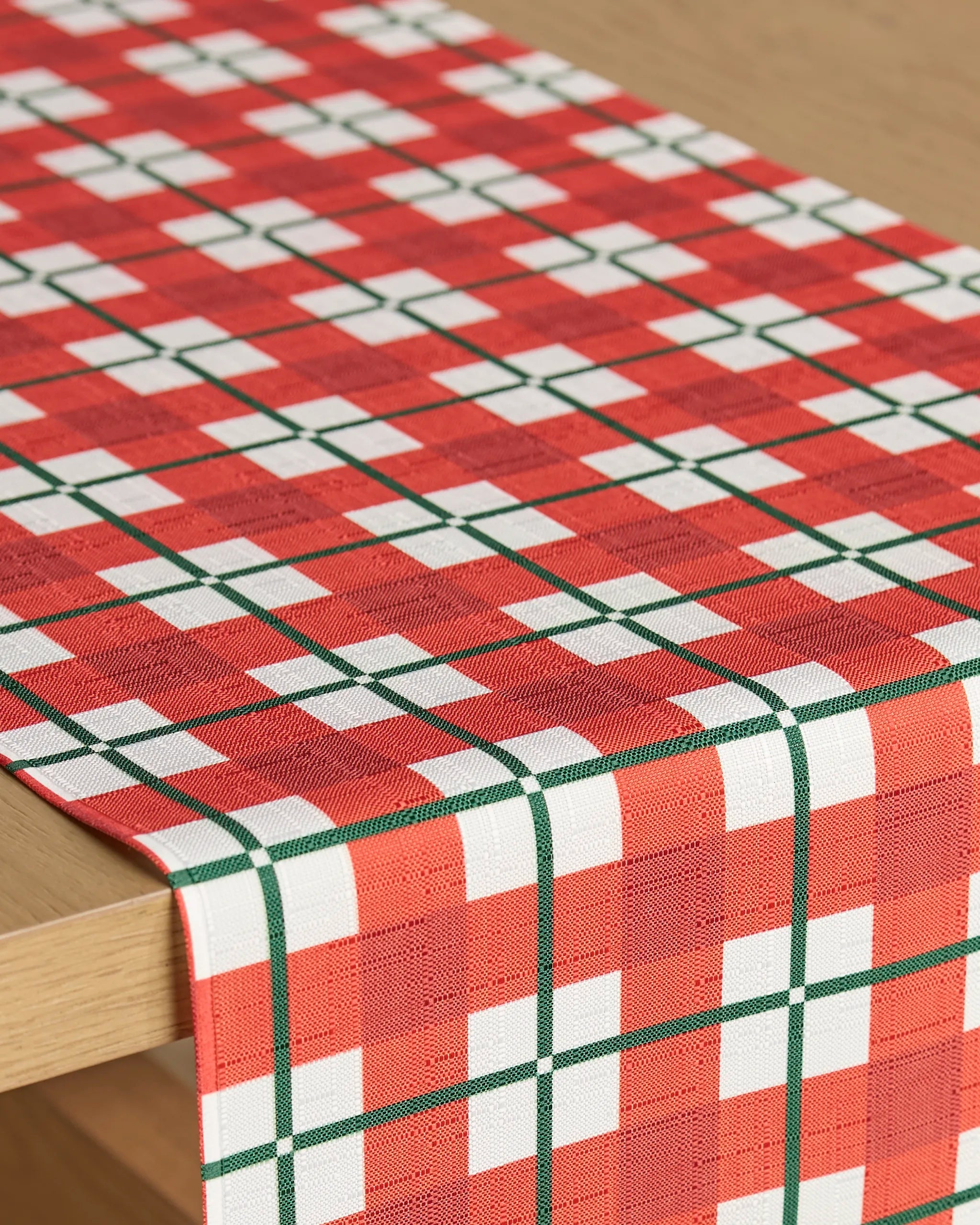 A close-up of the Geometry Yuletide Plaid stain-resistant table runner, showcasing its red, white, and green geometric checkered pattern on a light wooden table.