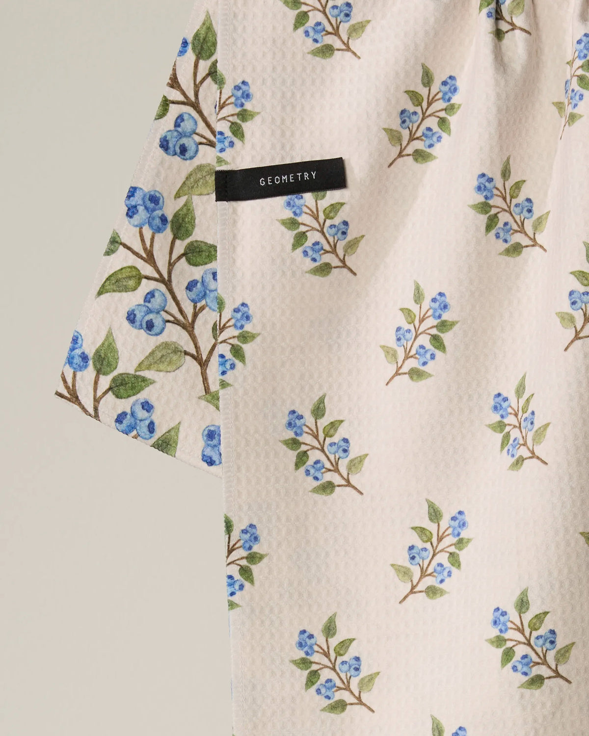 A close-up of the Blueberry Sprig tea towel reveals its textured white fabric adorned with blue and green floral sprigs. A black Geometry tag is attached, emphasizing this quick-drying towel's perfect blend of style and functionality.
