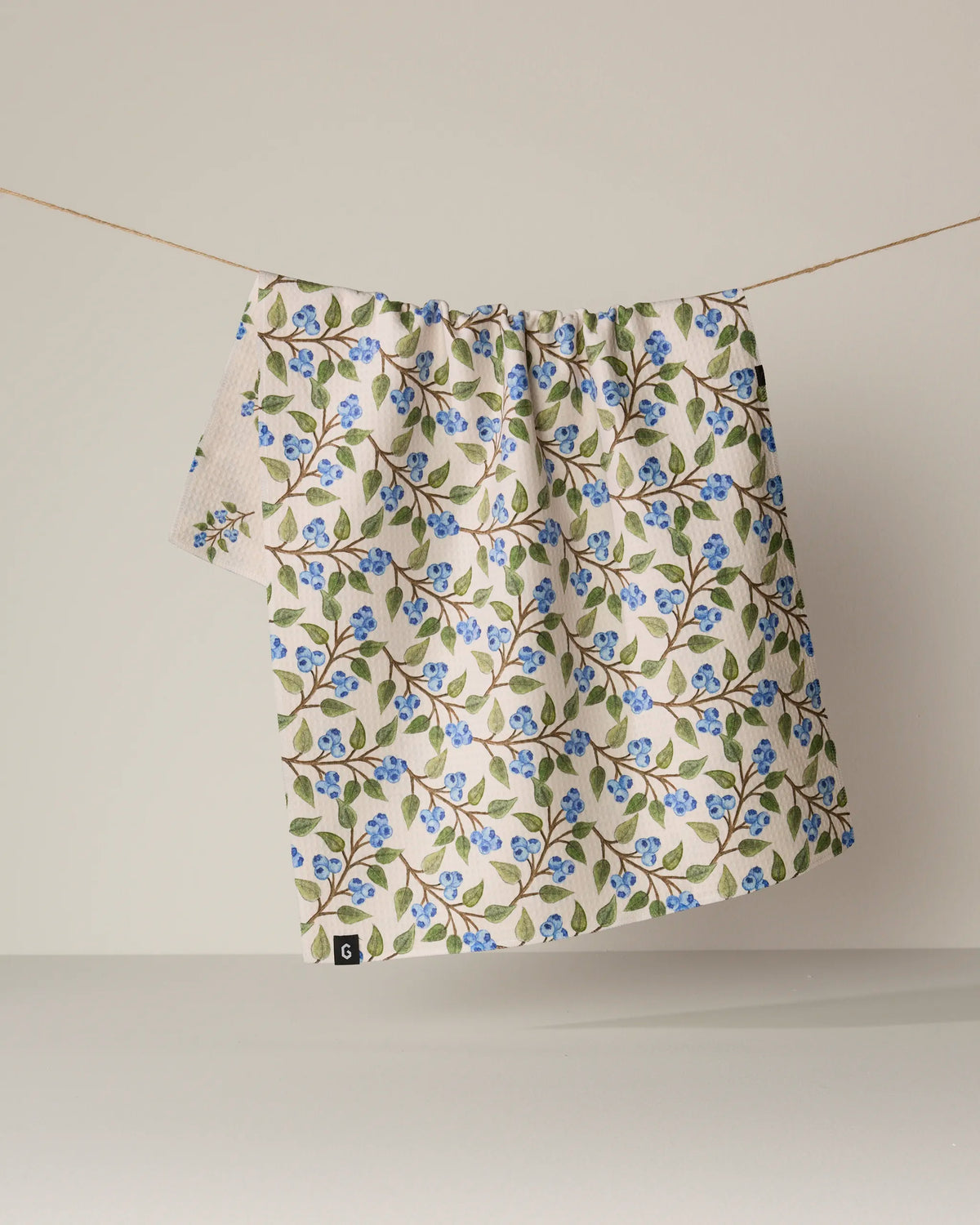 A cream-colored, double-sided tea towel from Geometry, featuring the "Blueberry Sprig" blue and green floral pattern, hangs on a string against a plain, light background.