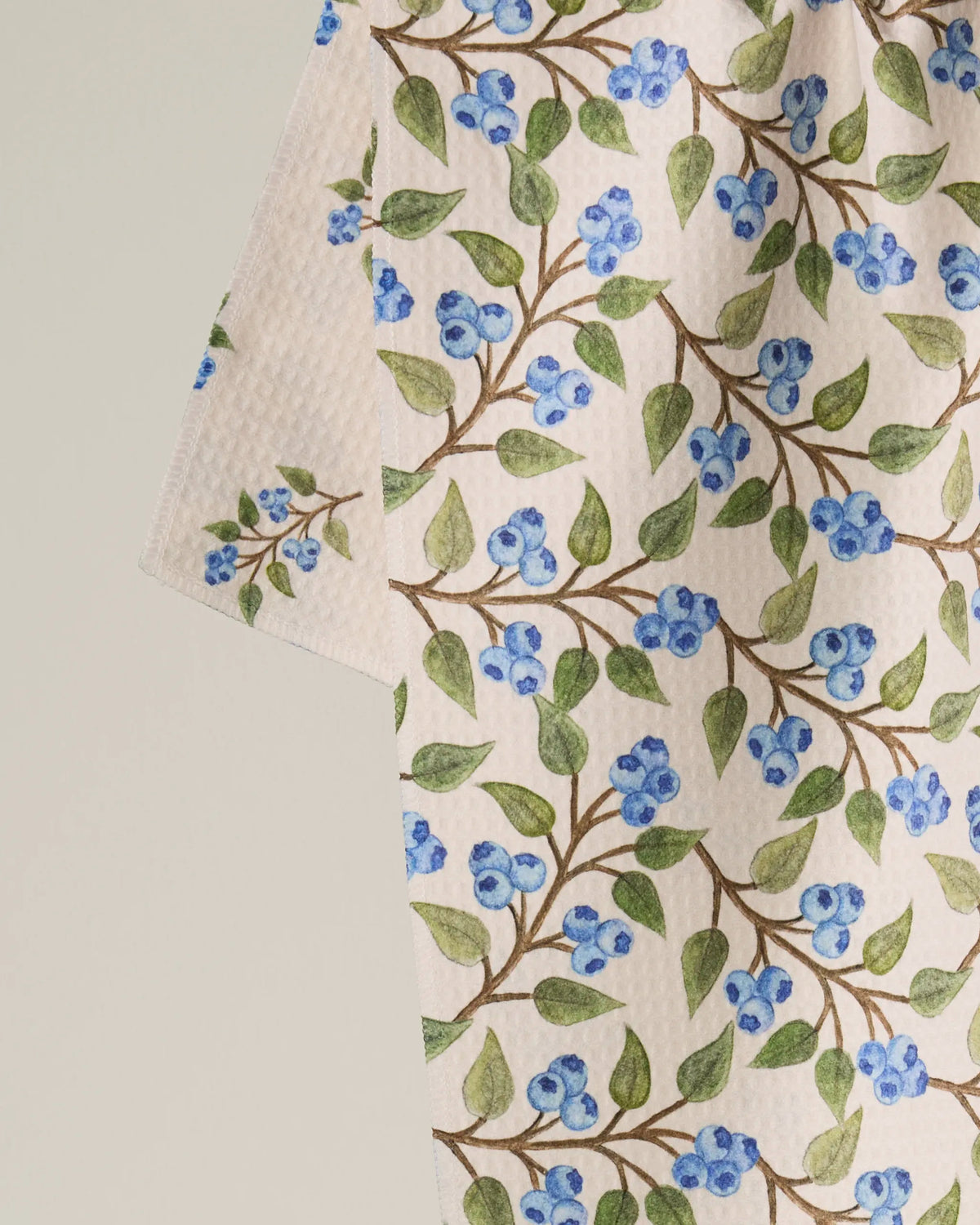 Introducing the Blueberry Sprig tea towel by Geometry—a double-sided design that showcases a crisp white background adorned with elegant green leaves and clusters of blue berries on delicate brown branches, presented against a simple light backdrop.