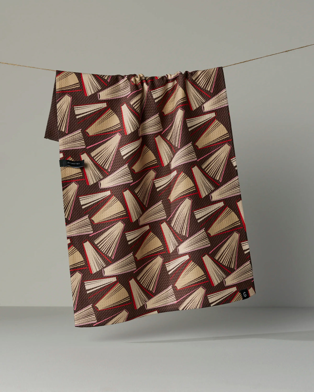 The Geometry “Bookmas” double-sided tea towel, featuring a geometric pattern of red, beige, and brown book illustrations, hangs on a thin string against a plain gray background.