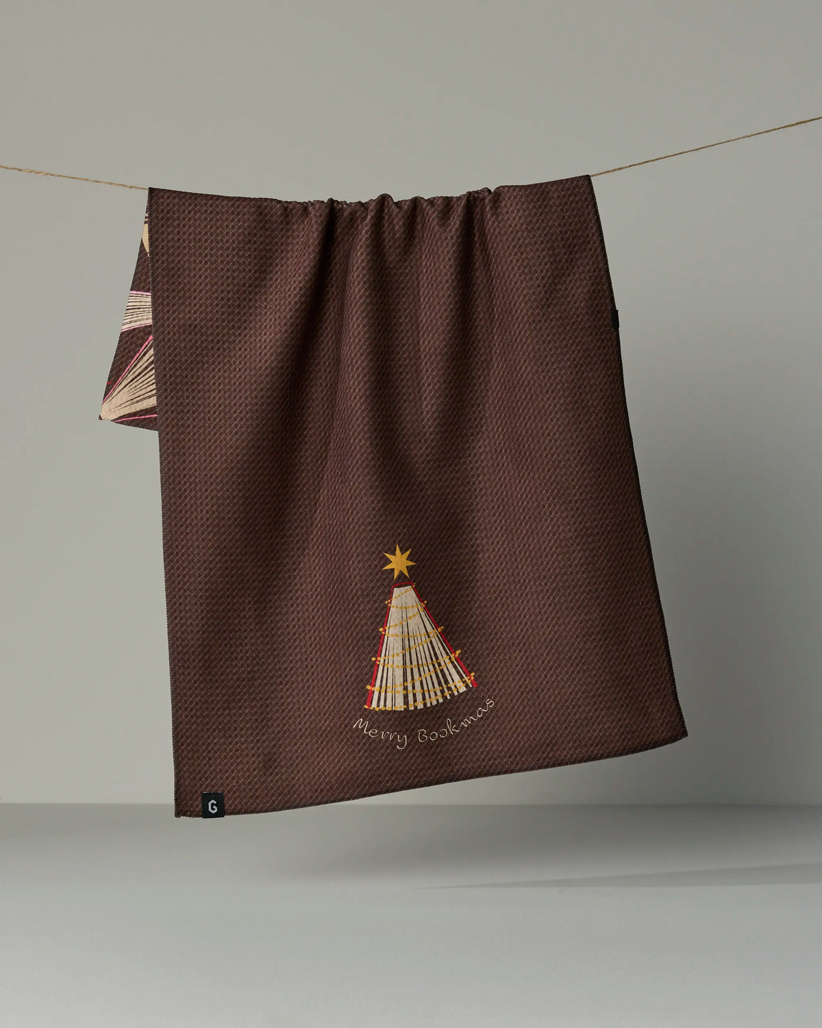 A brown textured recycled kitchen towel from Geometry, named “Bookmas,” hangs on a string, featuring an embroidered Christmas tree with a yellow star on top and the words “Merry Christmas” below.