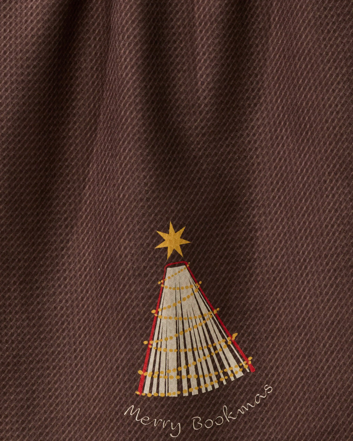 A festive scene featuring a tree crafted from stacked books topped with a yellow star, adorned like a Christmas tree, stands out on the brown textured background of Geometry’s recycled kitchen towel—capturing the Bookmas spirit, with “Merry Bookmas” displayed below.