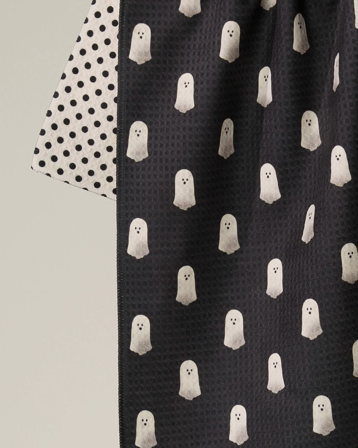 A close-up of the Geometry “Casper Boo” double-sided tea towel: one side features small black polka dots on a light background, while the other showcases white ghost illustrations on a dark background.