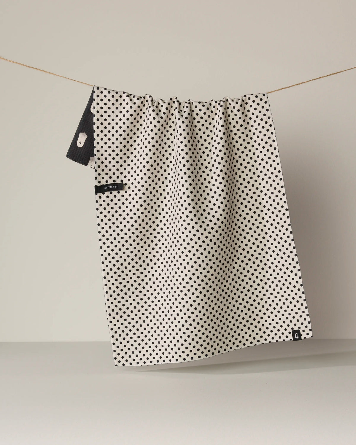 The Geometry Casper Boo tea towel, featuring a white quick-drying fabric with small black polka dots, hangs on a string against a plain background. A black label and a small loop are visible near the top on one side.