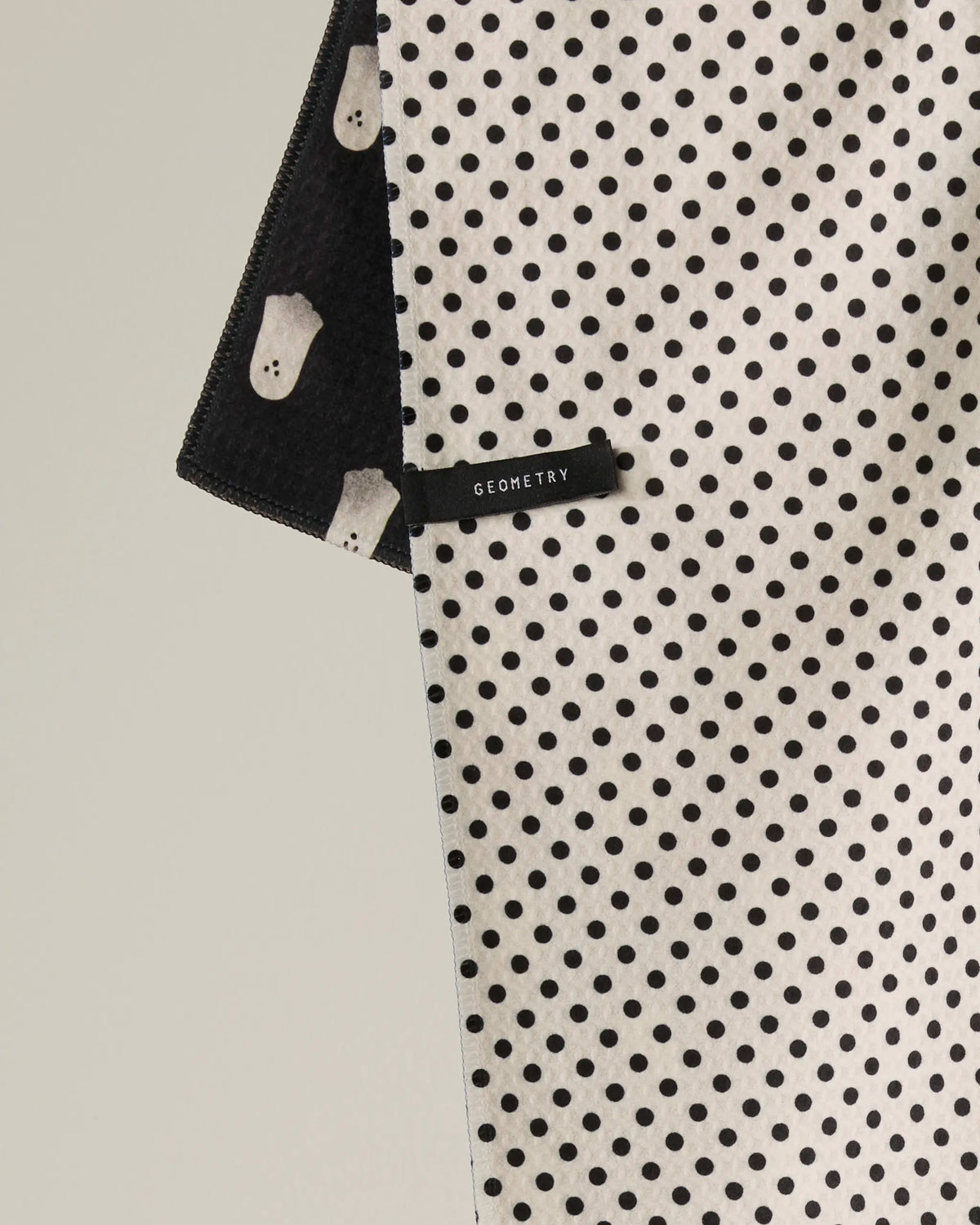 The Geometry Casper Boo tea towel features a black-and-white polka dot design with a visible Geometry label. The edge is folded to reveal the double-sided towel’s black underside, which is decorated with white ghost shapes, offering both style and functionality.