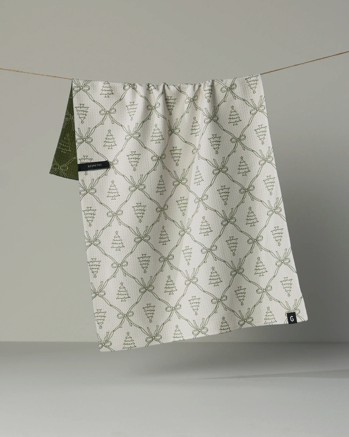 The Geometry “Cedar Bows” double-sided tea towel, featuring green botanical patterns, hangs on a string against a plain, light background—bringing both style and versatility to your kitchen.