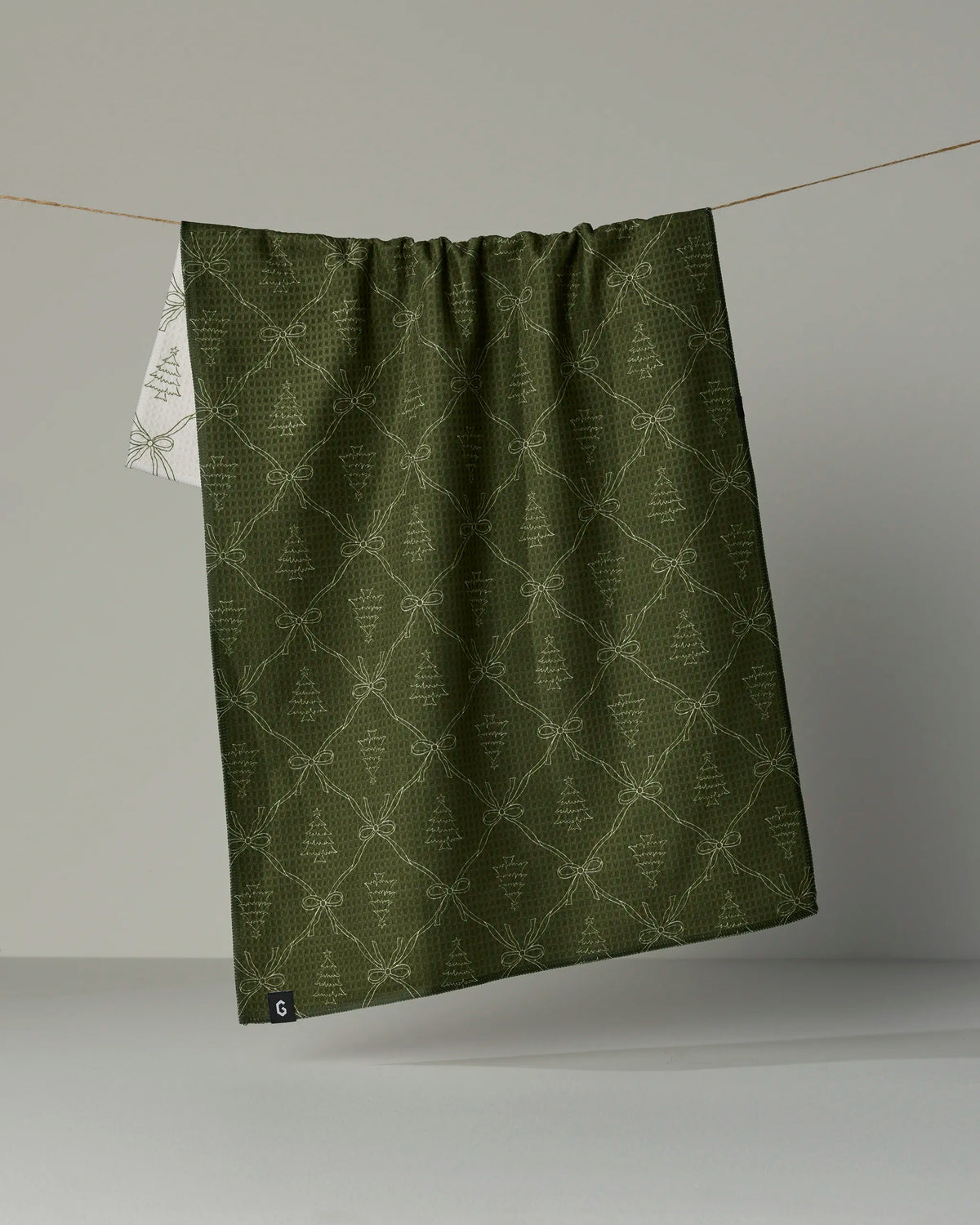 A green patterned Cedar Bows Double-Sided Tea Towel by Geometry hangs on a line against a plain gray background, with a small white tag visible in the lower left corner.