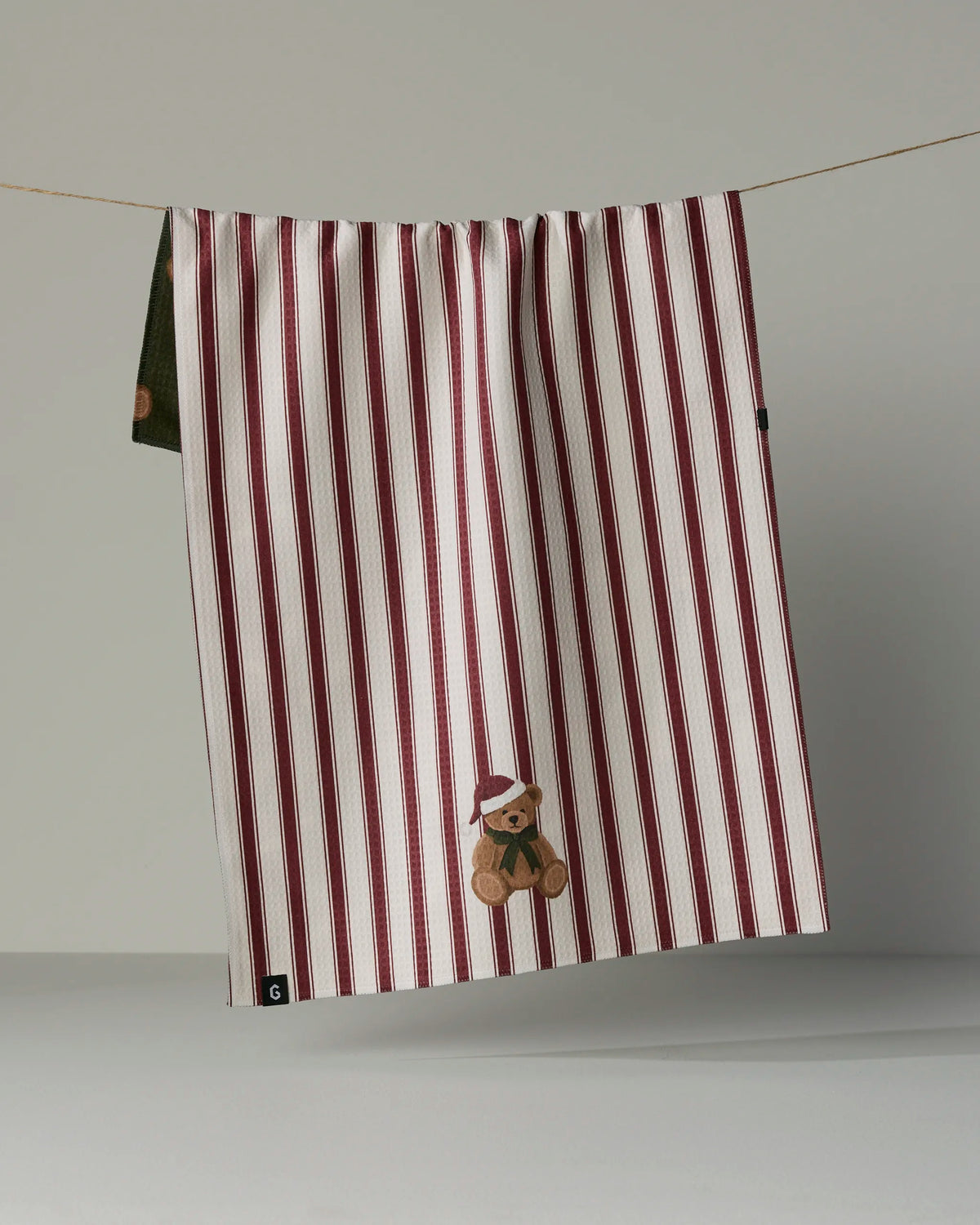 A white Geometry tea towel from the Christmas Bear collection, made from recycled materials, hangs on a line. It features vertical burgundy stripes and an embroidered teddy bear in a red beret near the bottom corner.