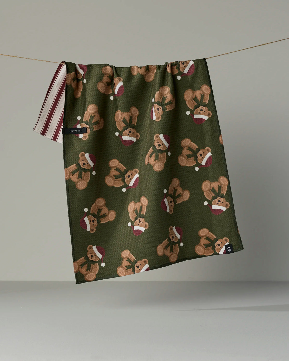 The Geometry Christmas Bear quick-drying towel, in dark green and patterned with brown teddy bears wearing red Santa hats, hangs on a line. This double-sided tea towel features striped trim on one side and brand tags in the bottom corner.