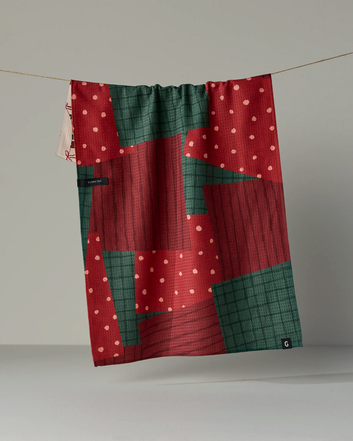 The Geometry “Christmas Crafts” tea towel, featuring red and green double-sided designs with pink polka dots and plaid patterns, hangs on a string against a plain light gray background. Crafted from recycled materials, this quick-drying towel blends festive style with sustainable quality.