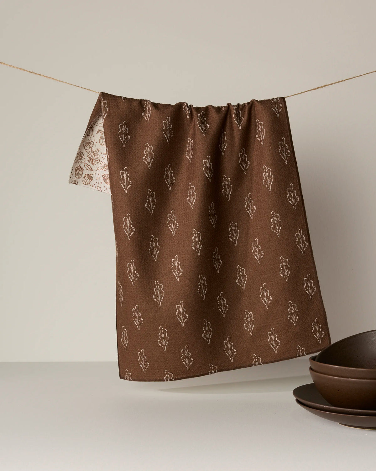 A double-sided Cozy Flora tea towel by Geometry, featuring an earthy brown pattern with white leaf designs, hangs on a string. Crafted from recycled materials, its reverse side displays a lighter coordinating motif. Brown ceramic bowls are arranged on the plain neutral surface below.