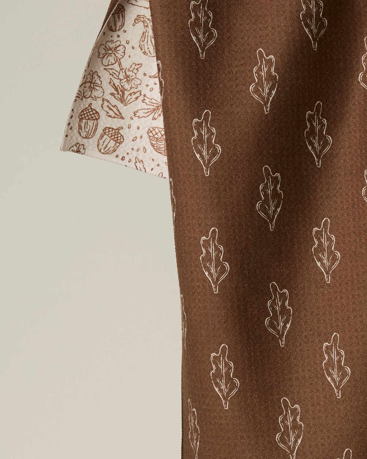 Introducing the Cozy Flora tea towel by Geometry. This double-sided towel features a rich brown fabric adorned with a delicate white oak leaf pattern on one side, while the edge reveals a cream lining decorated with acorns, leaves, and flowers in subtle light brown outlines. Perfect for adding style to your kitchen, the Cozy Flora is crafted from recycled materials for an eco-friendly touch.