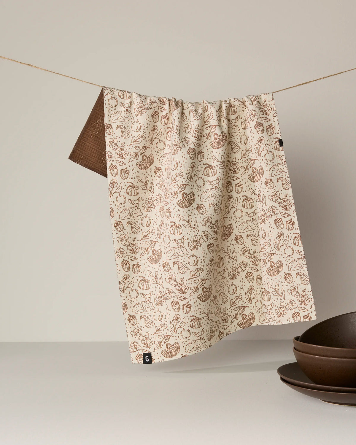 A beige, double-sided tea towel from Geometry’s Cozy Flora collection, featuring a brown vegetable pattern and made from recycled materials, hangs on a string. Nearby, brown bowls are stacked on a white surface against a simple light background.