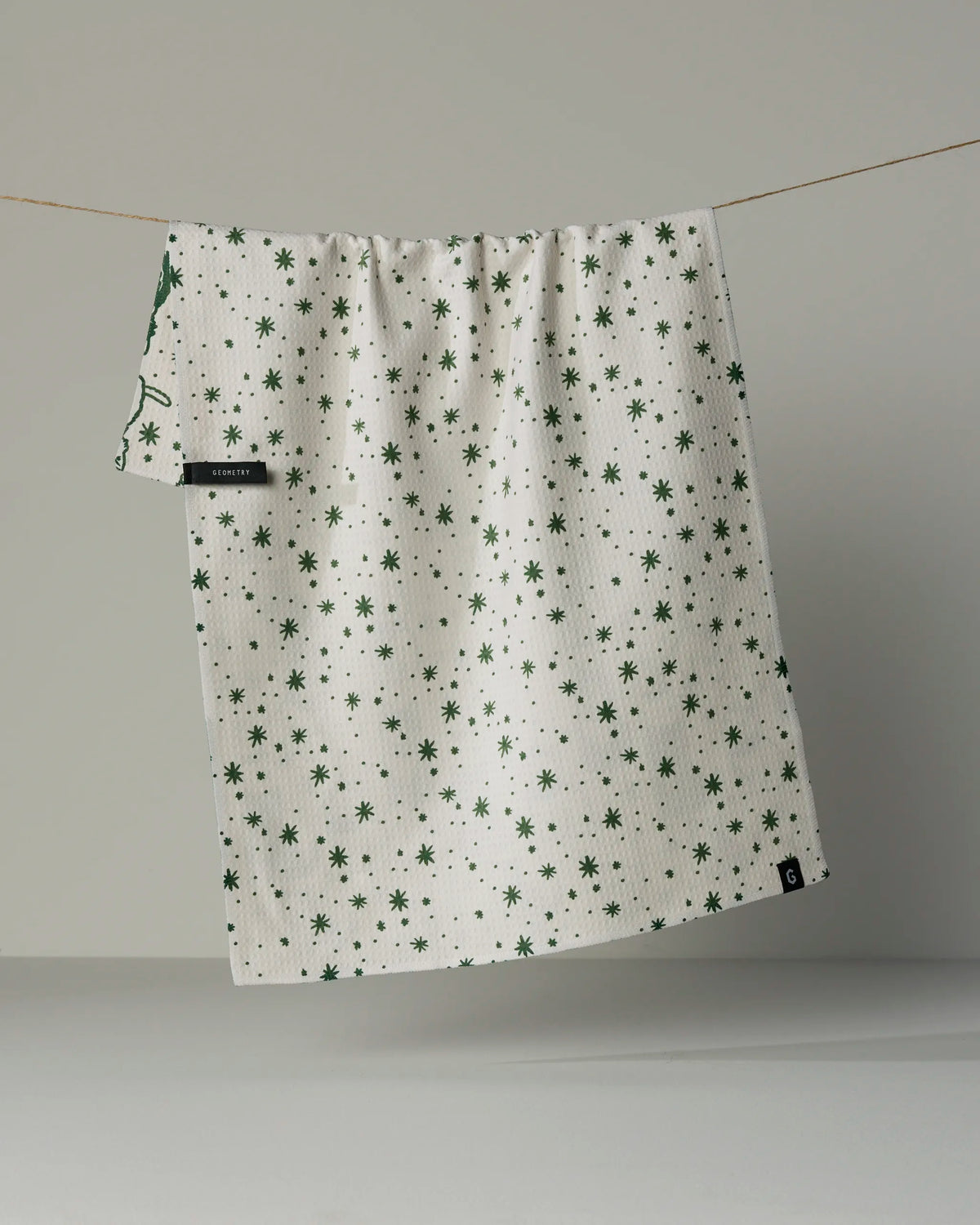 A Geometry “Cozy Kittens” tea towel, featuring a white background with a green starburst pattern and made from quick-drying material, hangs on a clothesline against a plain gray backdrop.
