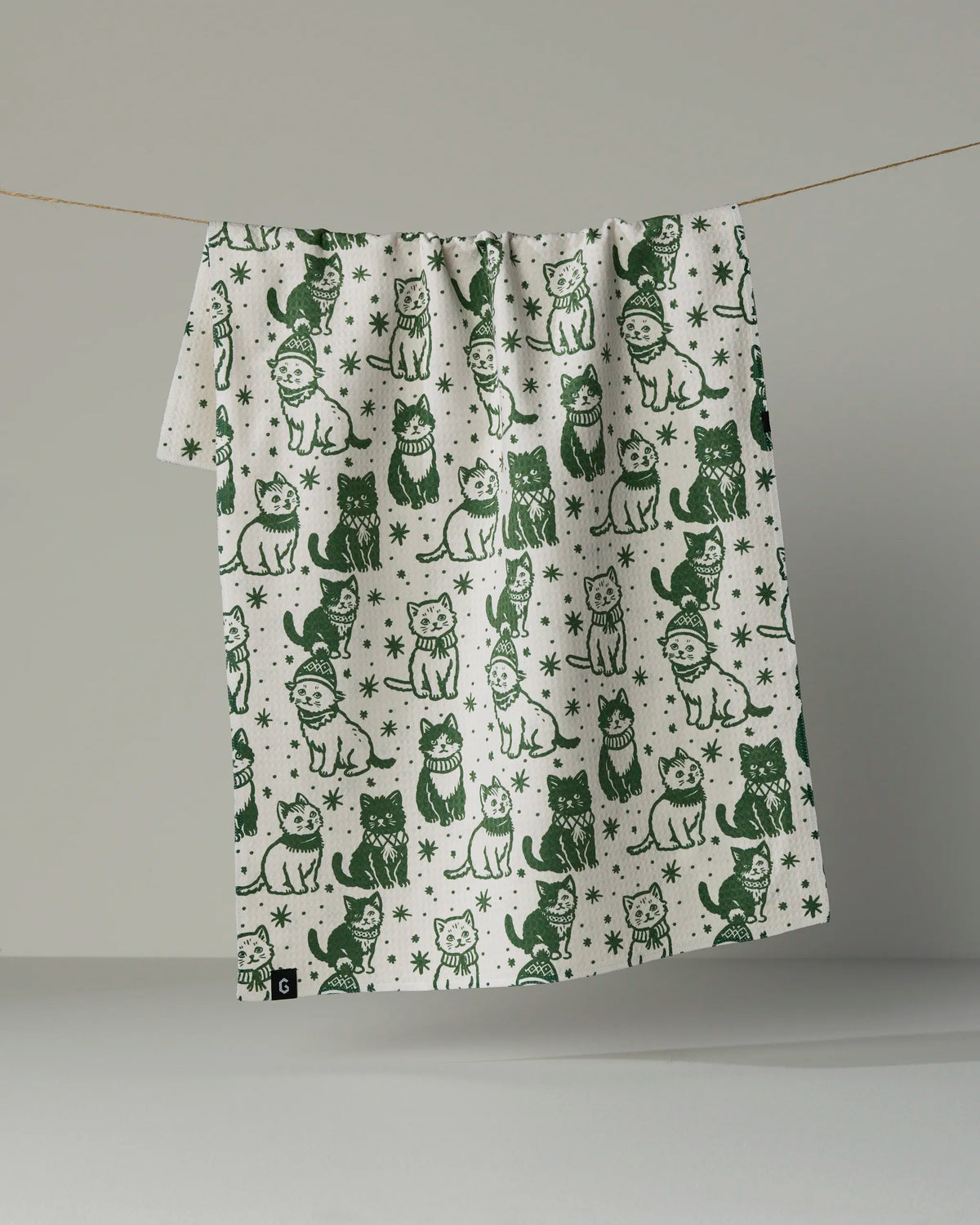 The Cozy Kittens tea towel by Geometry, featuring green illustrations of playful and festive cats in hats and scarves surrounded by stars and snowflakes on a white, quick-drying fabric, hangs on a string against a plain background.