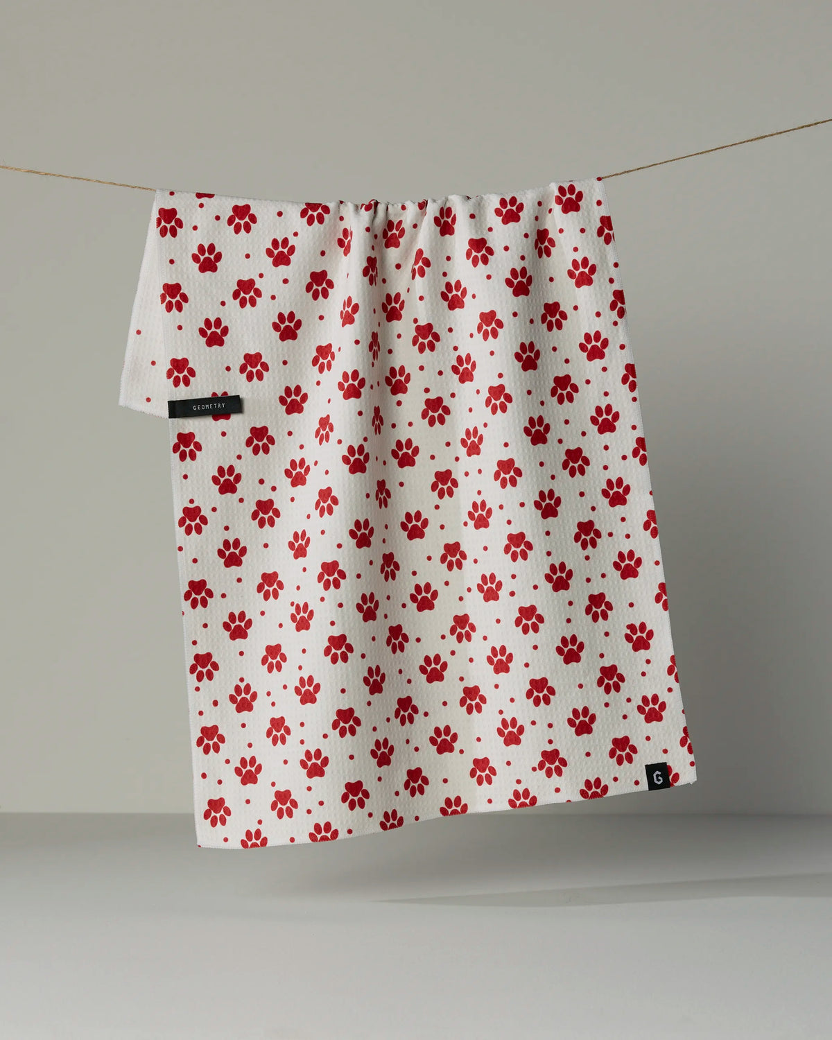 A white Geometry Dachshund Dash Kitchen Tea Towel featuring a pattern of red paw prints hangs on a string against a plain gray background, crafted from sustainable GeoLUXE Fabric.