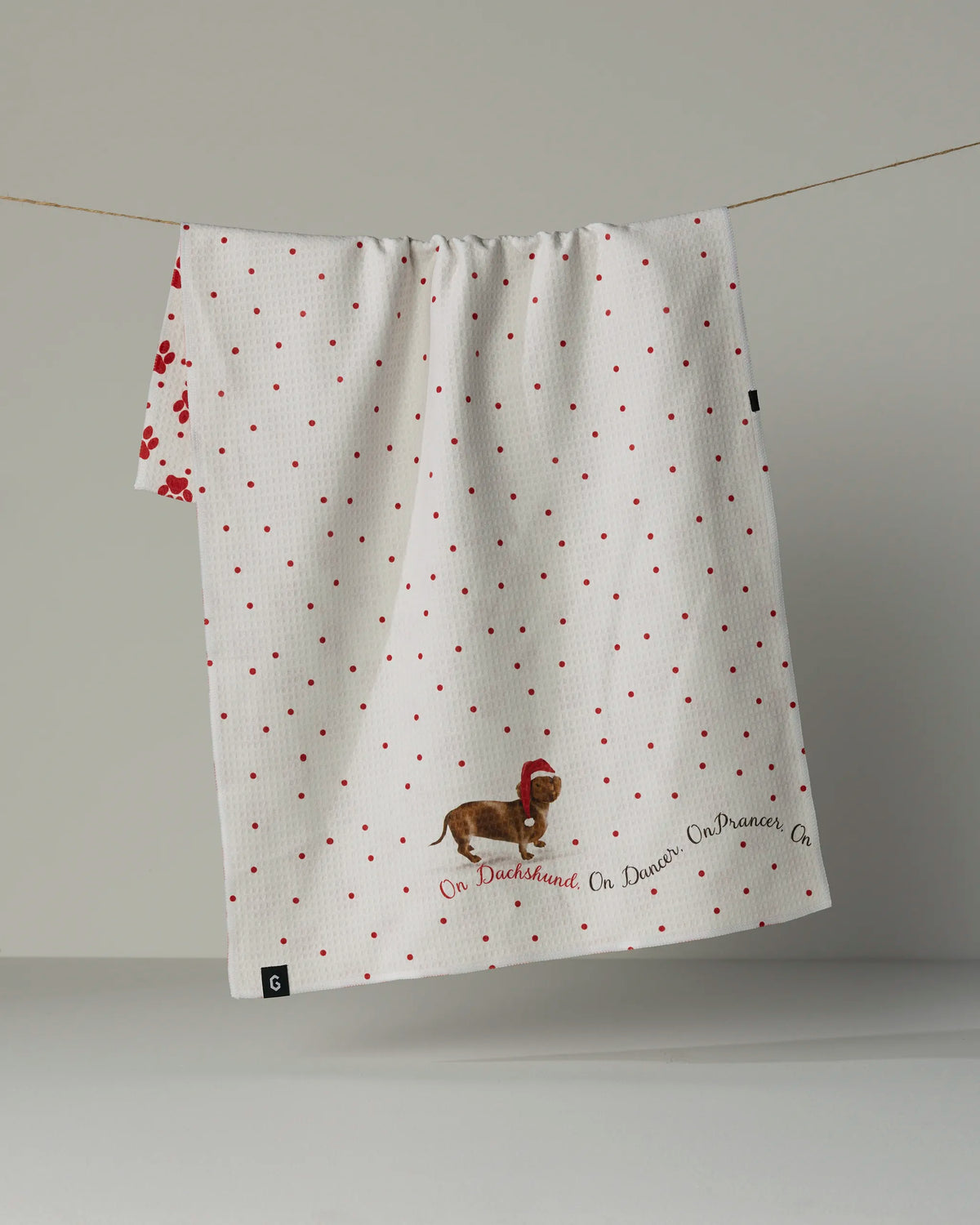 A white towel from Geometry, the Dachshund Dash, hangs on a line. Featuring an illustration of a dachshund wearing a red hat and the playful text “On Dachshund, On Dancer, On Prancer,” this kitchen tea towel is crafted from recycled materials with premium GeoLUXE Fabric—bringing both charm and sustainability to your kitchen décor.