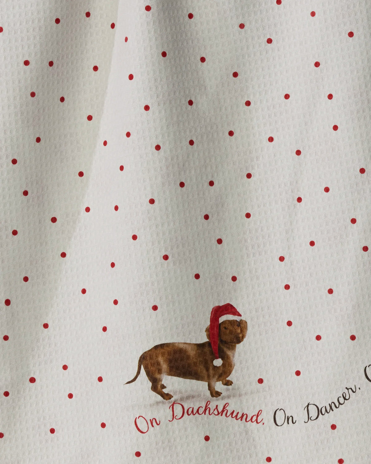 A white Geometry “Dachshund Dash” Kitchen Tea Towel showcases a charming illustration of a dachshund in a red Santa hat against playful red polka dots. Crafted from recycled GeoLUXE fabric, this festive towel features the vibrant red cursive phrase, “On Dachshund, On Dancer.”.