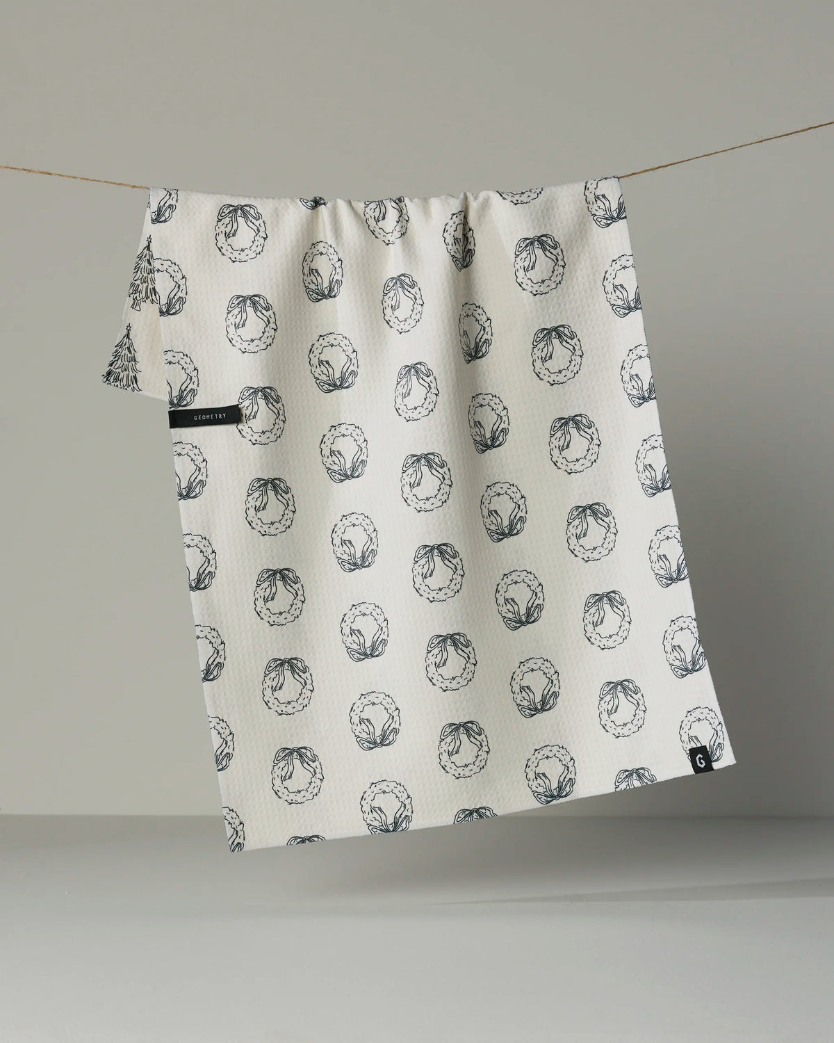 A white, quick-drying tea towel from Geometry called “Evergreen Sketches,” featuring a repeating black line art pattern, hangs on a string against a plain background.