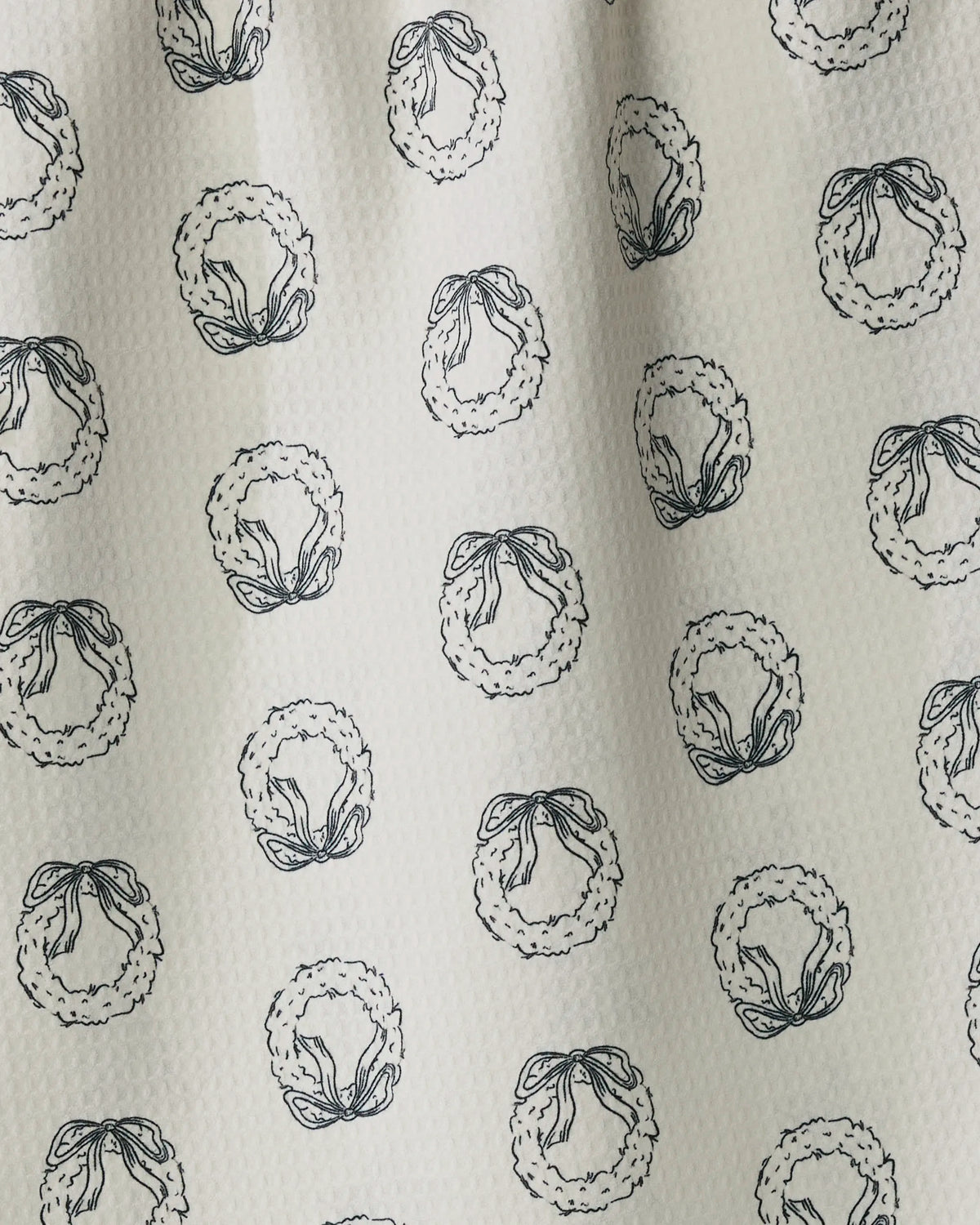 Evergreen Sketches by Geometry features a white textured fabric adorned with a repeating pattern of hand-drawn wreaths tied with bows, each outlined in black and evenly spaced across the surface. This quick-drying tea towel combines style and functionality, making it ideal for everyday use in the kitchen.