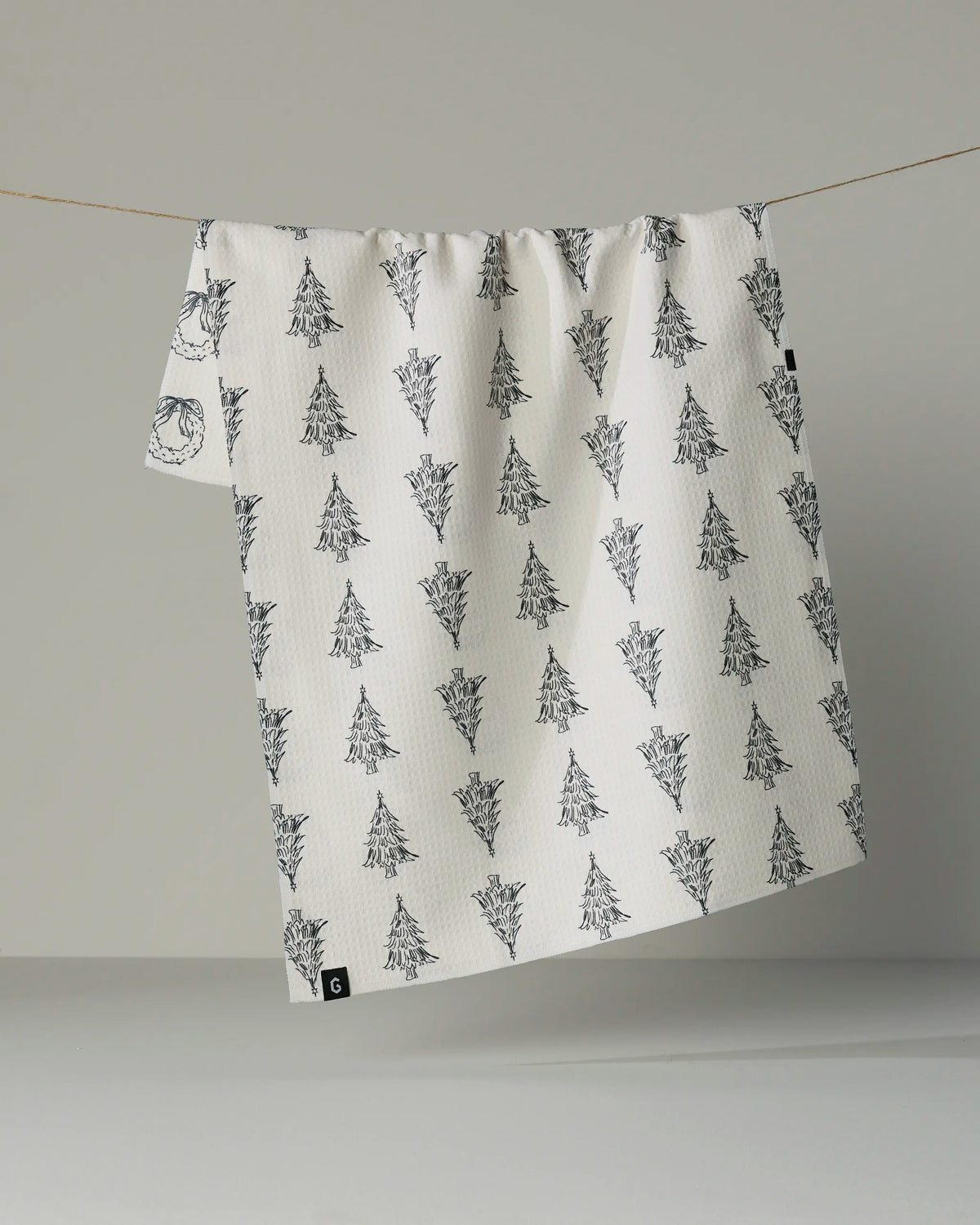 The Geometry “Evergreen Sketches” double-sided tea towel, in crisp white and adorned with a repeating pattern of black-inked pine trees, hangs on a clothesline against a plain gray background.