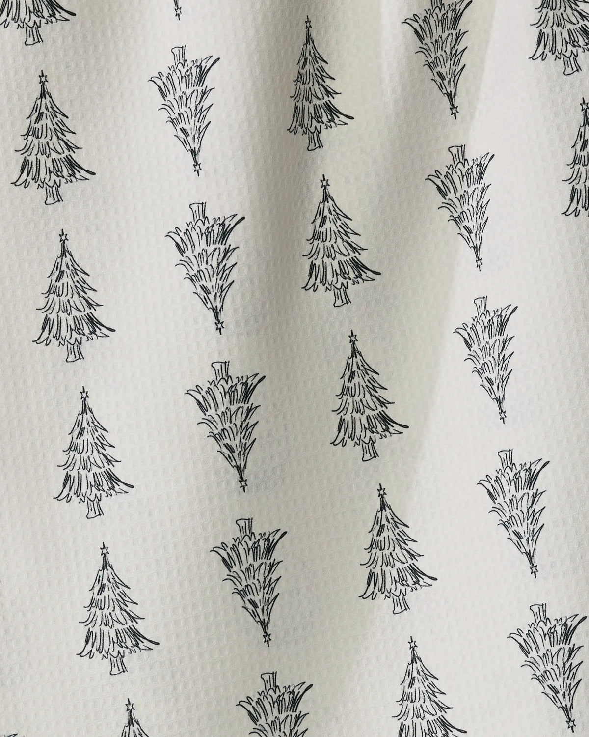 Evergreen Sketches by Geometry features a white textured fabric adorned with a repeating pattern of black, hand-drawn Christmas trees arranged in crisp rows and columns—some standing upright, others playfully upside down. Subtle shadows and gentle wrinkles add natural character to the surface of this quick-drying tea towel, making it an ideal way to bring festive charm to your kitchen.