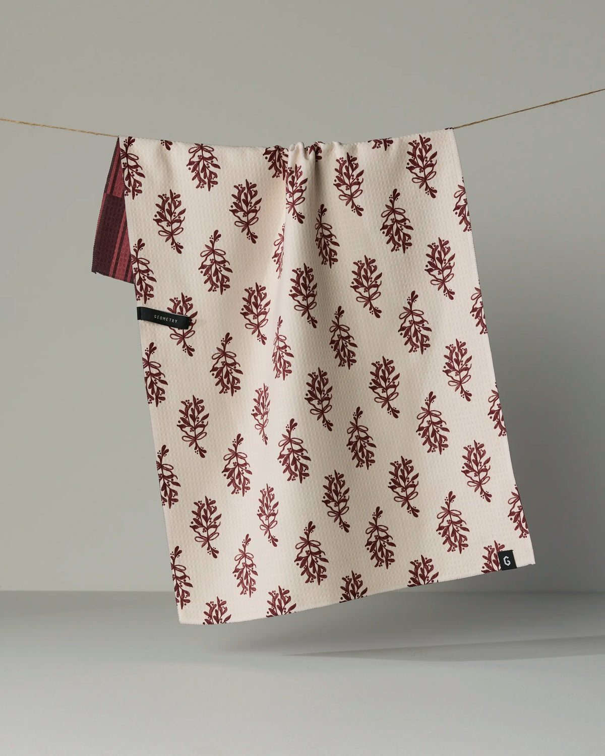 The Geometry “Festive Cranberry” double-sided tea towel, crafted from recycled materials, features a red leafy pattern on beige and hangs on a clothesline against a gray background. Quick-drying and adorned with a small black tag in one corner.