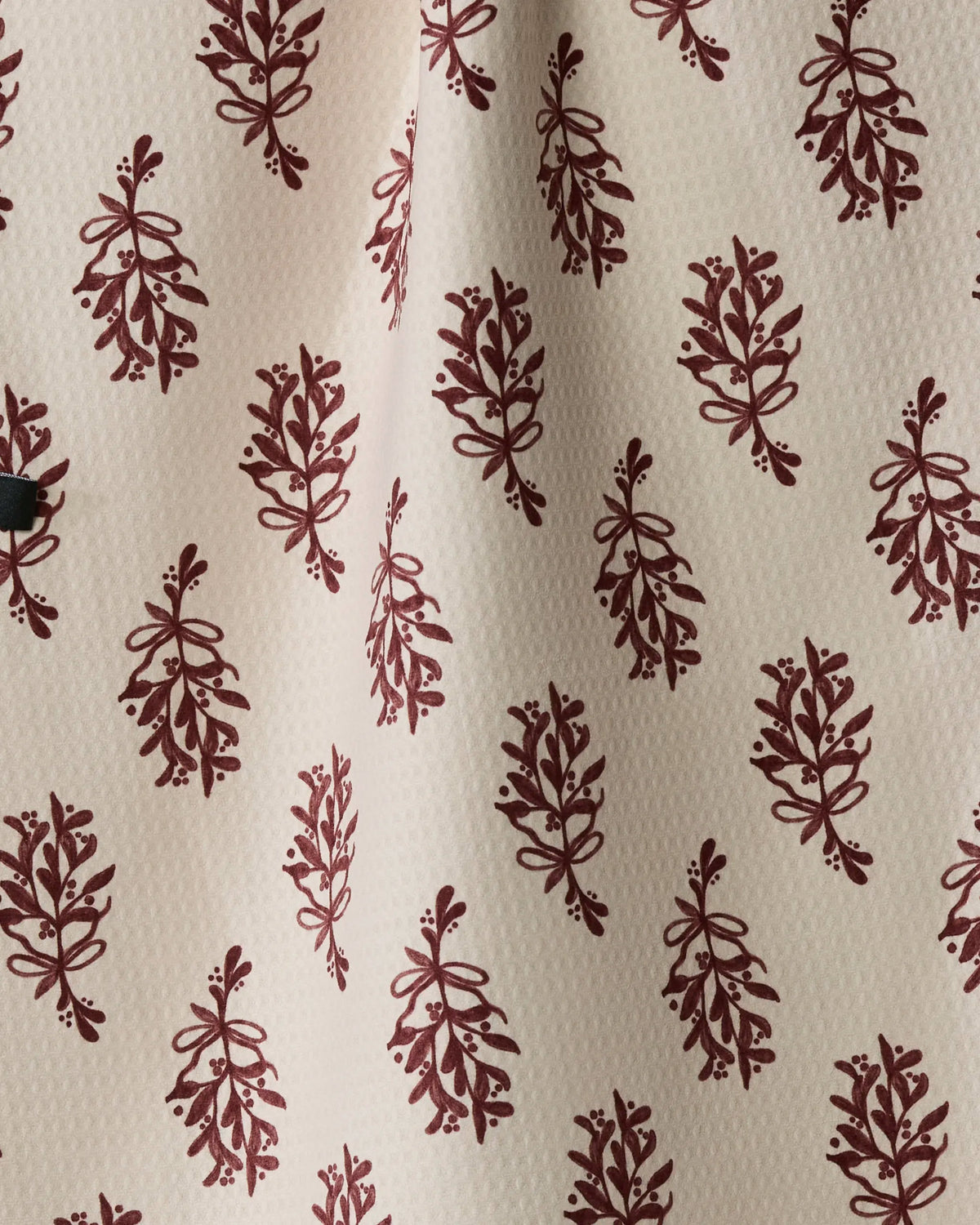 Introducing the “Festive Cranberry” tea towel by Geometry: crafted from recycled materials, this double-sided kitchen accessory features a white fabric with a textured surface and a repeating maroon botanical motif arranged in neat, evenly spaced rows. Boasting quick-drying properties, it includes a subtle vertical fold through the material.