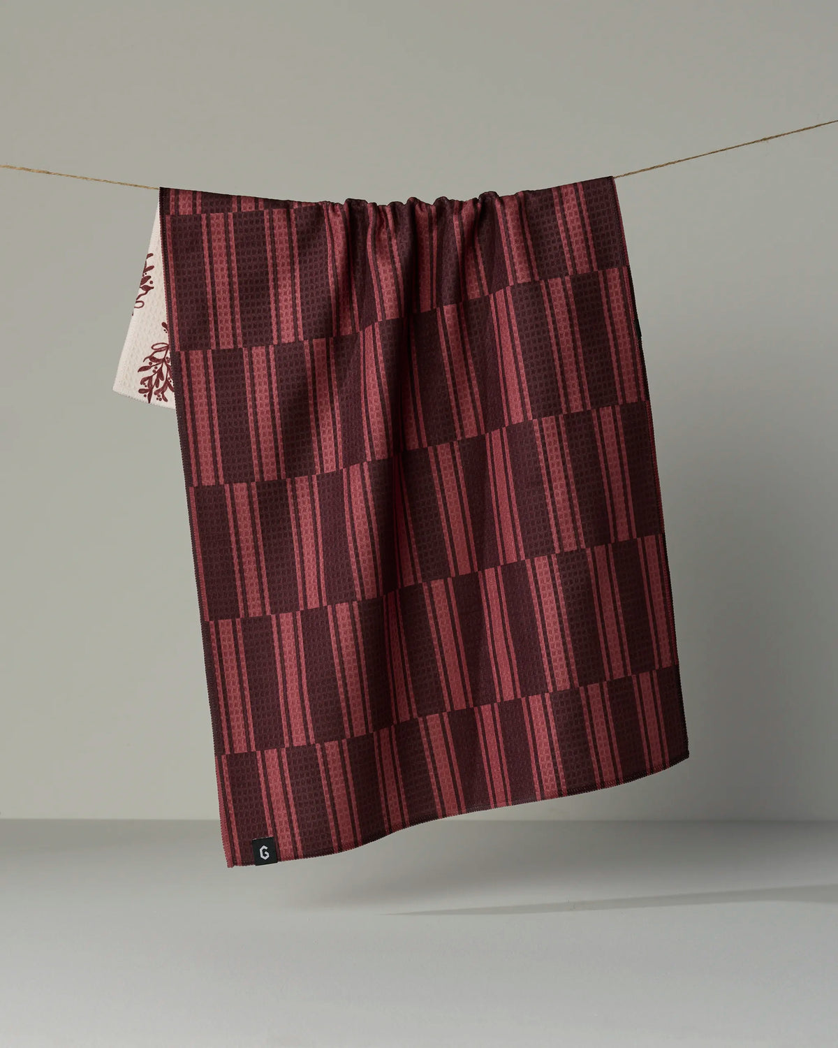 A Geometry Festive Cranberry tea towel, featuring a maroon and dark red checkered pattern and crafted from recycled materials, hangs on a string against a plain gray background, with a small corner of a white patterned double-sided towel visible on the left side.