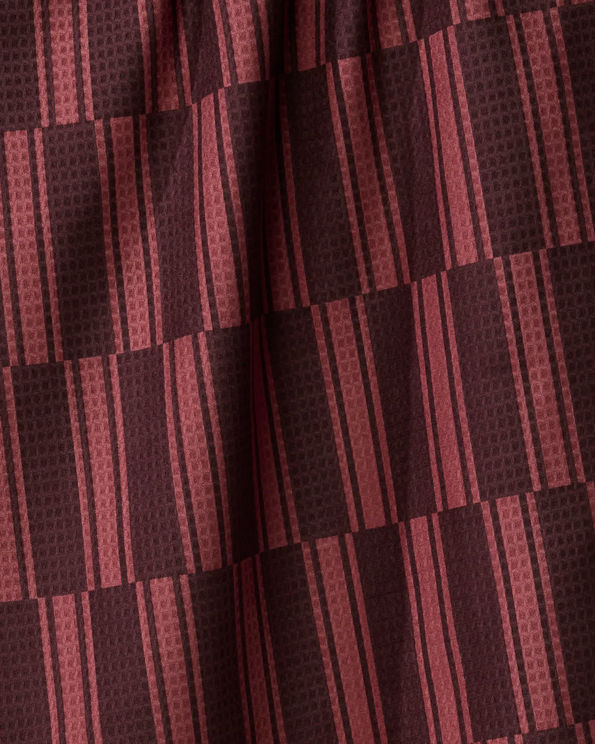 A close-up of the Festive Cranberry tea towel by Geometry showcases recycled fabric with a geometric pattern of alternating dark brown and cranberry-red vertical and horizontal stripes, creating a plaid or checkered effect. The double-sided design displays subtle folds and textured details.