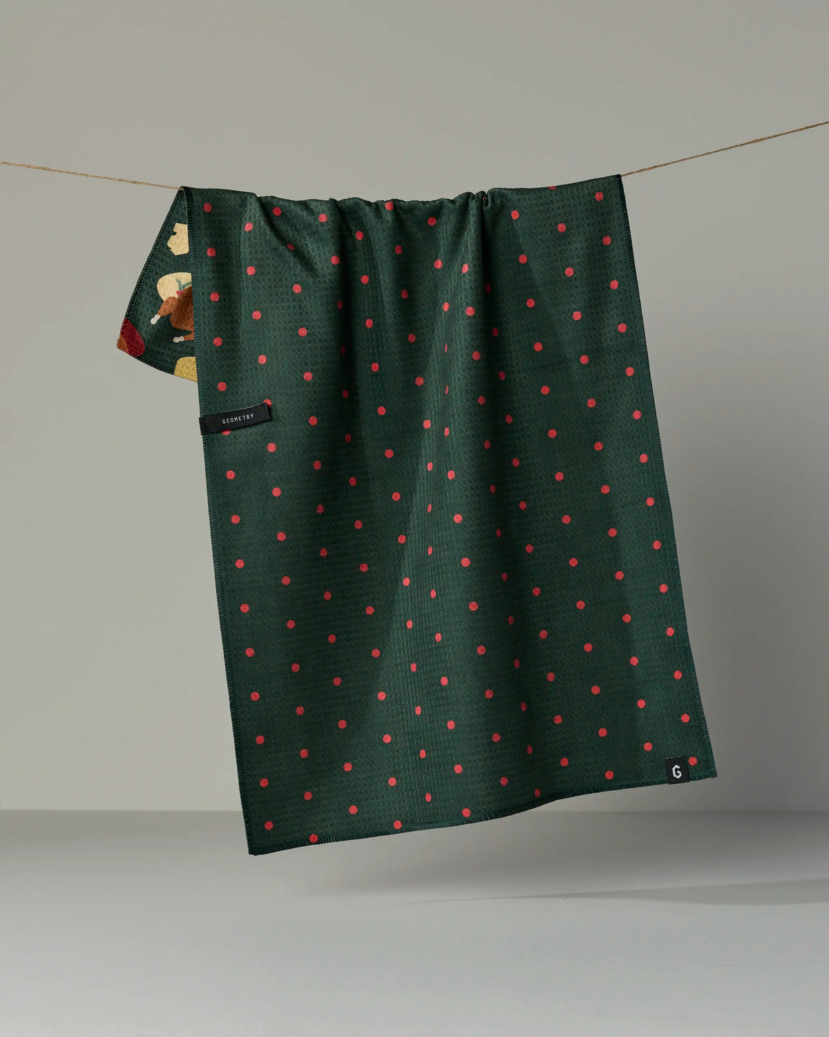 A dark green double-sided tea towel from Geometry, called Festive Feast, with small red polka dots hangs on a line. A corner reveals a hidden floral pattern on the reverse side. A black label and a small white Geometry logo tag are visible on the front.