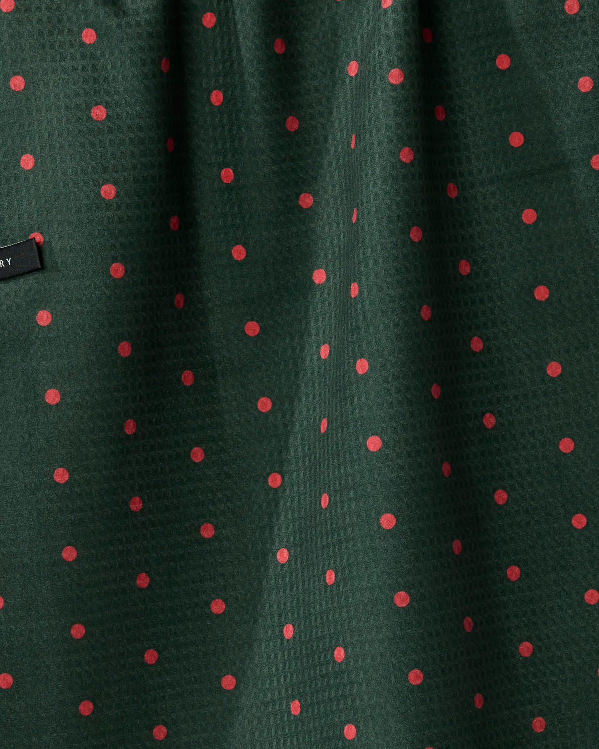 Crafted by Geometry, the Festive Feast tea towel showcases a dark green fabric adorned with a subtle textured pattern and evenly spaced small red polka dots. Designed for quick drying, it also features a partially visible small black fabric tag on the left side of the image.