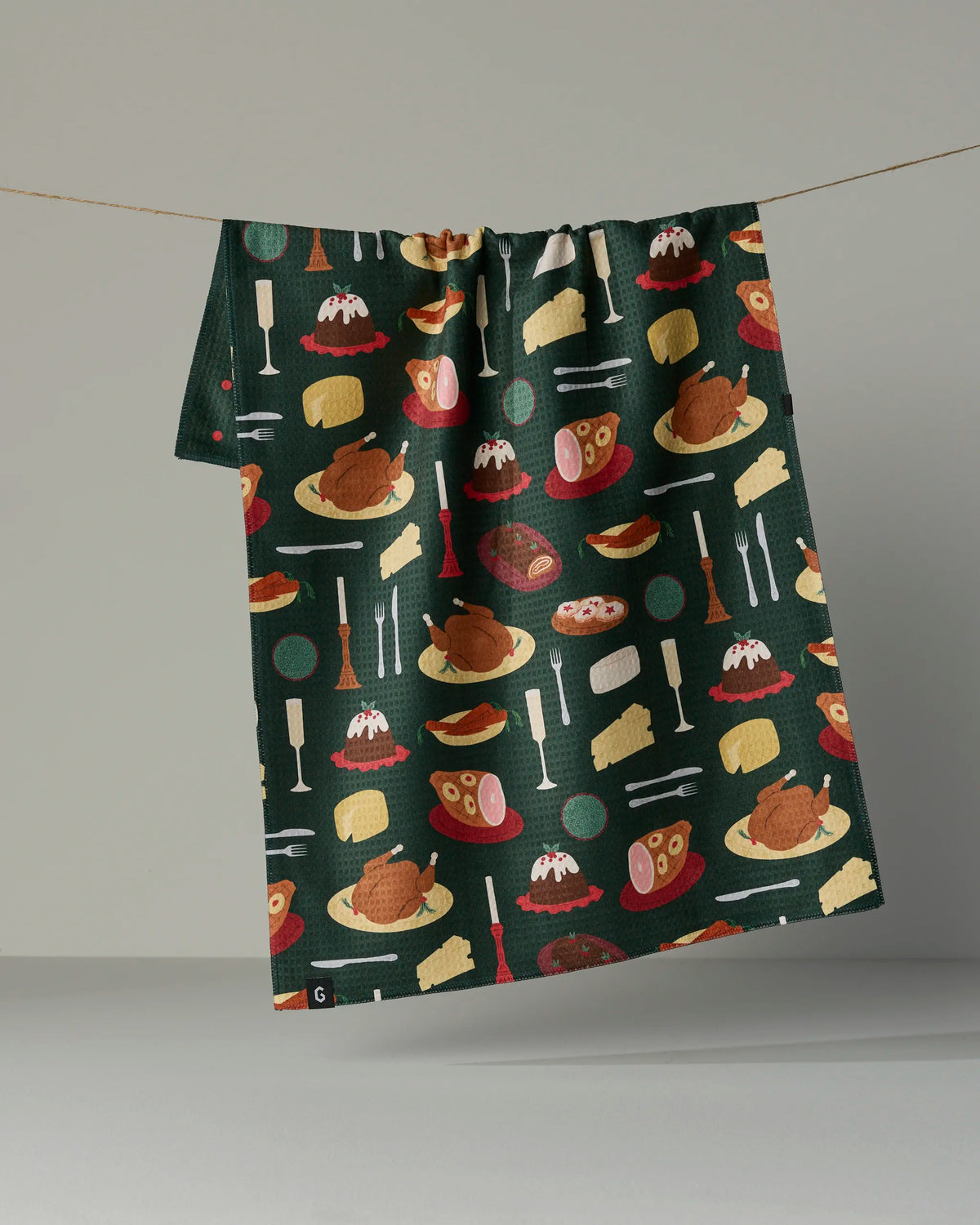 A dark green, quick-drying Festive Feast tea towel by Geometry hangs on a line, adorned with colorful illustrations of assorted food dishes, utensils, and drinks in a repeating pattern.