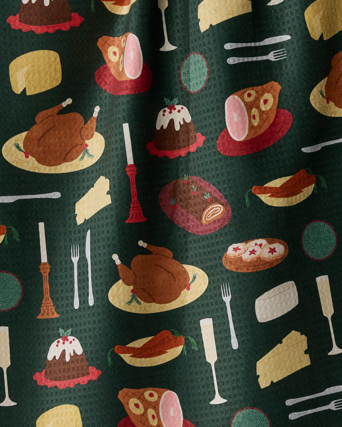 The Geometry Festive Feast double-sided tea towel features a patterned fabric adorned with illustrations of roast chicken, ham, cheese, carrots, desserts, plates, forks, knives, wine glasses, and candlesticks set against a dark green background.