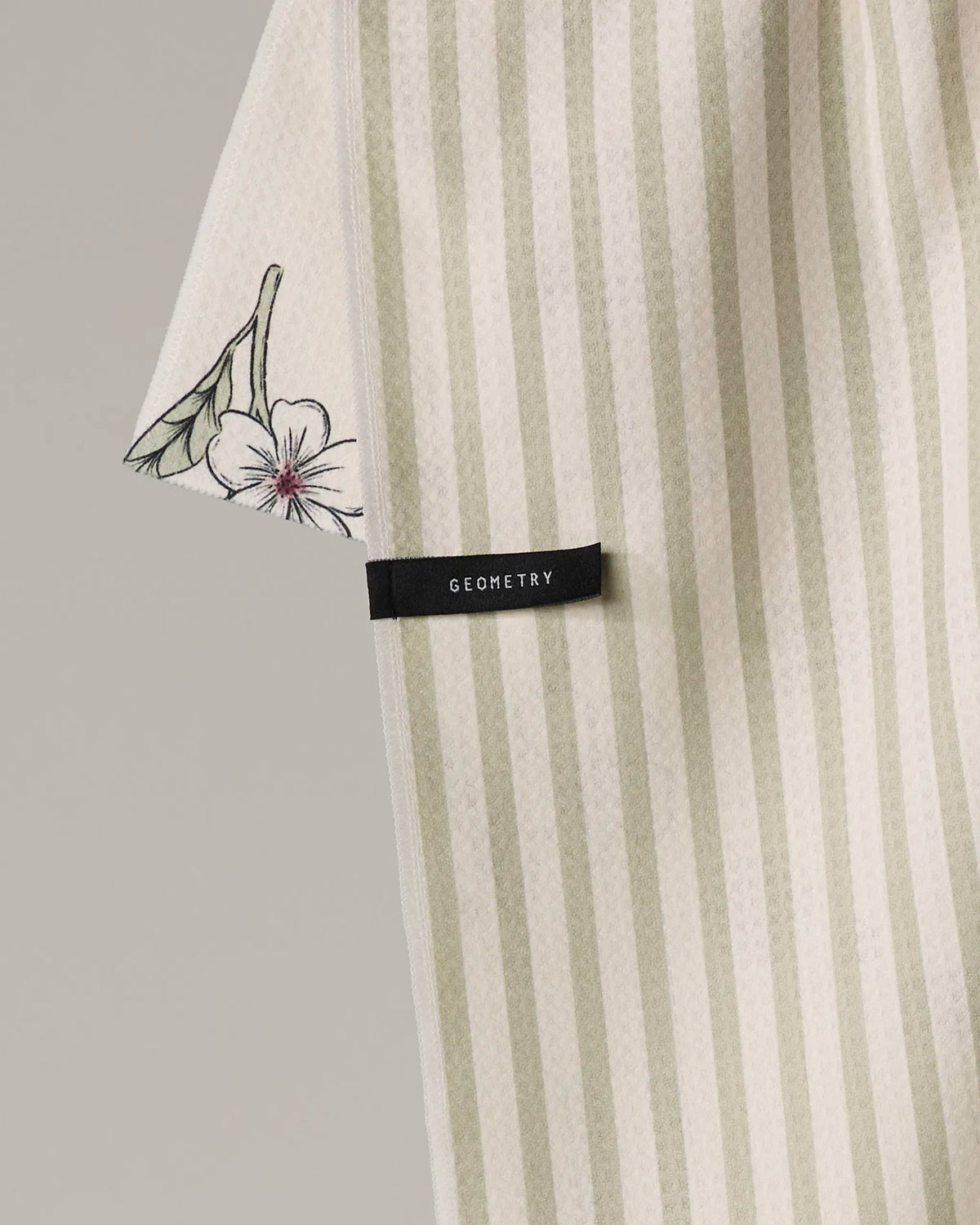 A Garden of Fortune Geometry towel featuring sage green and cream vertical stripes, a floral print on one corner, and a small black brand label.