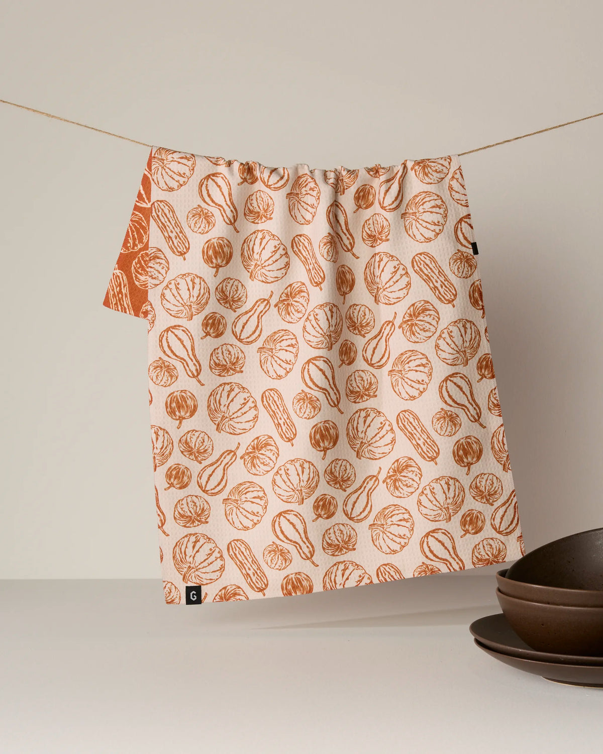 The Geometry Harvest Sketch double-sided tea towel—featuring an orange pumpkin and squash pattern on beige and sustainably crafted from recycled materials—hangs on a string, while brown ceramic bowls are stacked nearby on a light surface.