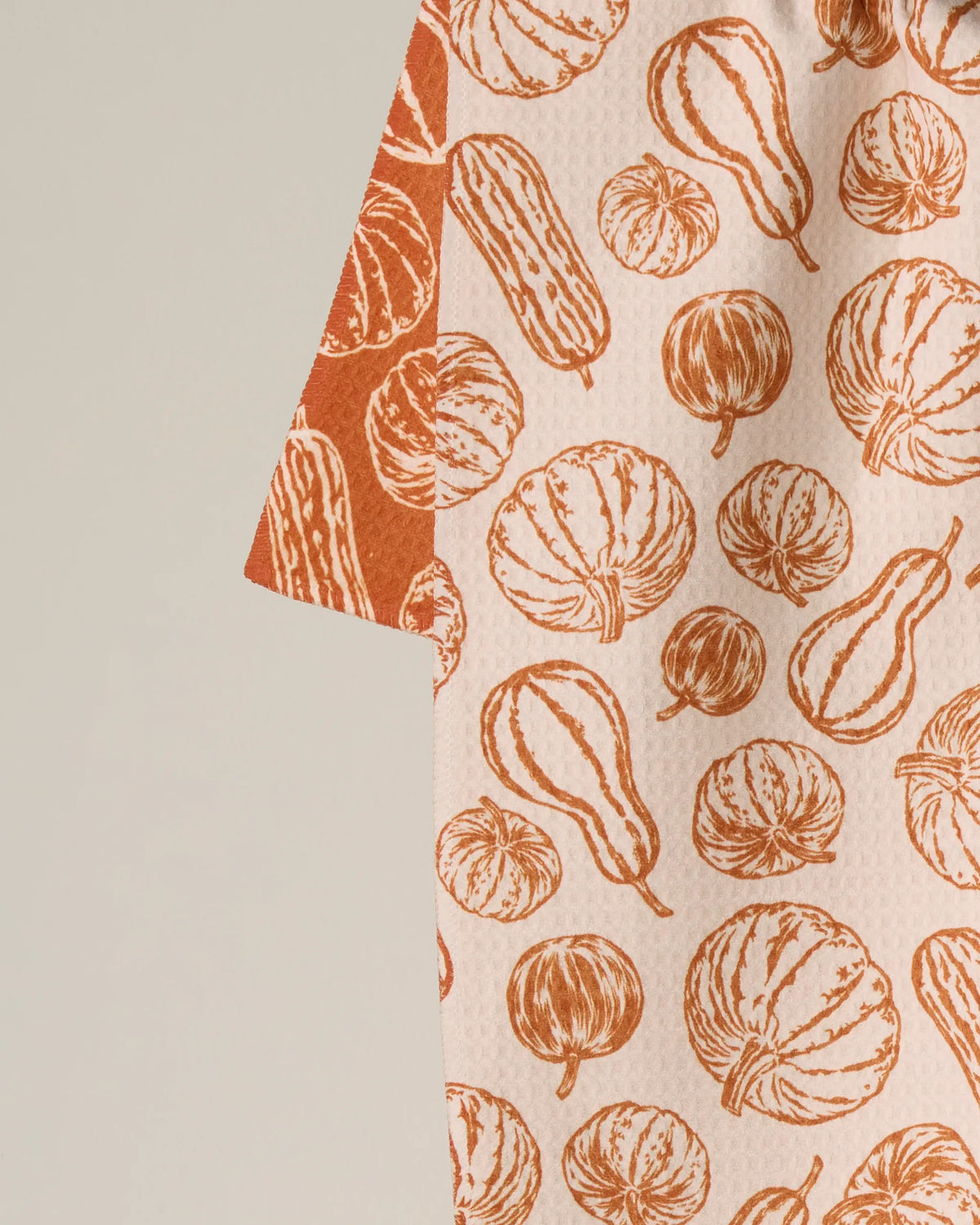 A close-up of the Geometry Harvest Sketch tea towel showcases an orange pumpkin and gourd pattern on a cream background. The edge features a contrasting darker orange section with the same harvest-inspired design, making this double-sided towel both stylish and versatile. Crafted from recycled materials, it brings an eco-friendly touch to your kitchen while celebrating seasonal charm.